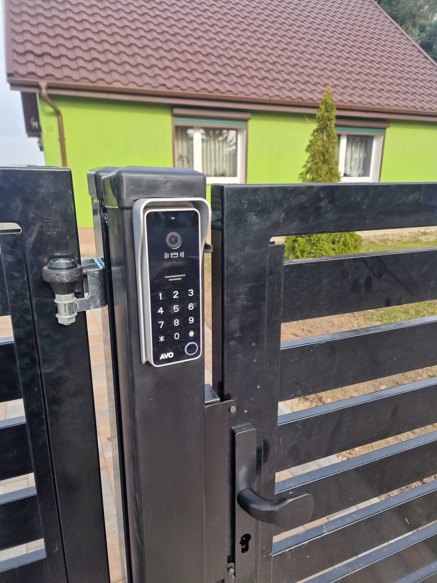 Nowoczesny wideodomofon z klawiaturą numeryczną zamontowany na czarnej, poziomej bramie wjazdowej; w tle zielony dom z brązowym dachem.