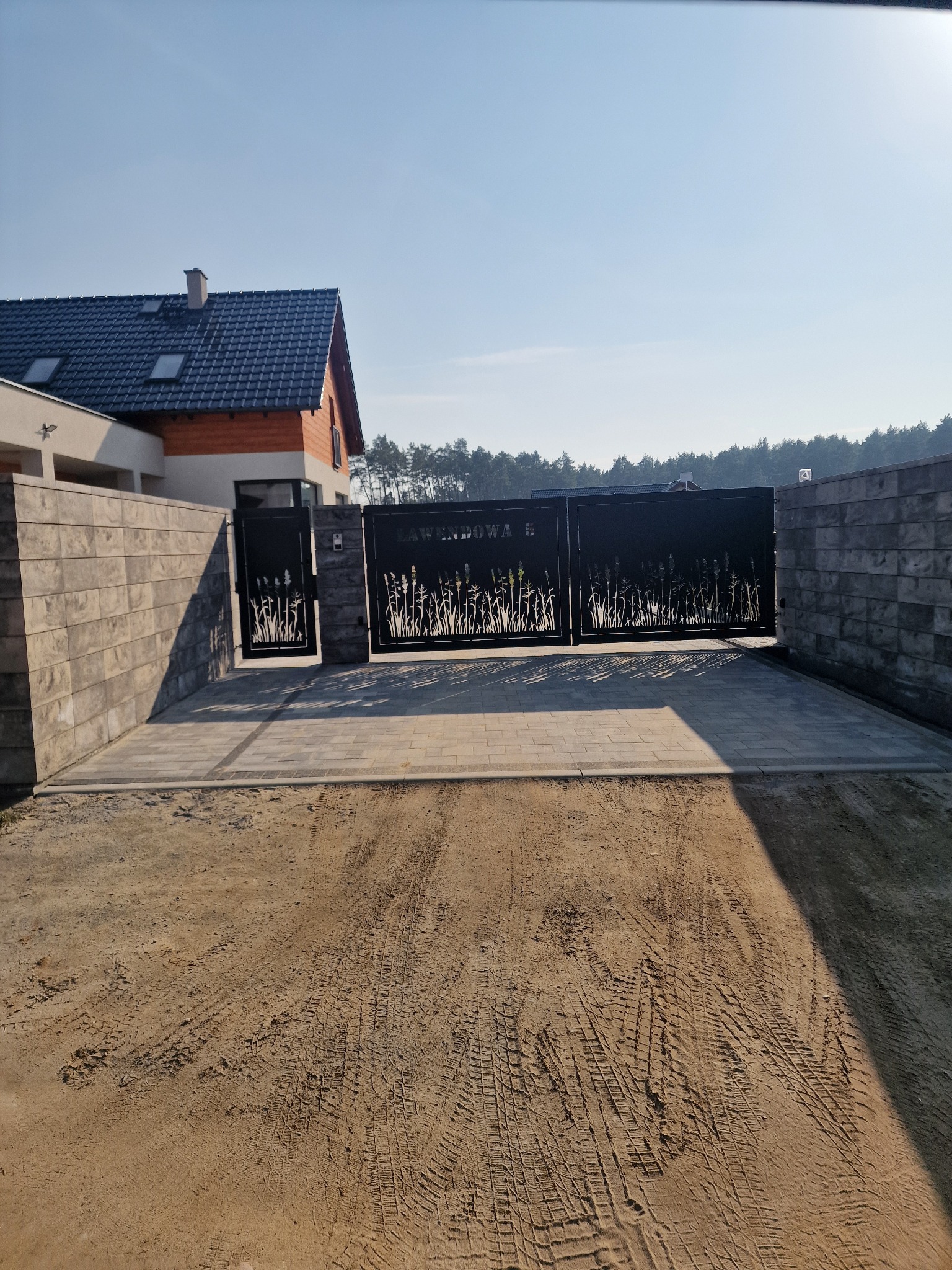 Nowoczesna brama wjazdowa z metaloplastyki z motywem roślinnym, zamontowana na posesji z kostki brukowej i szarego muru, widoczne ślady opon na piasku.