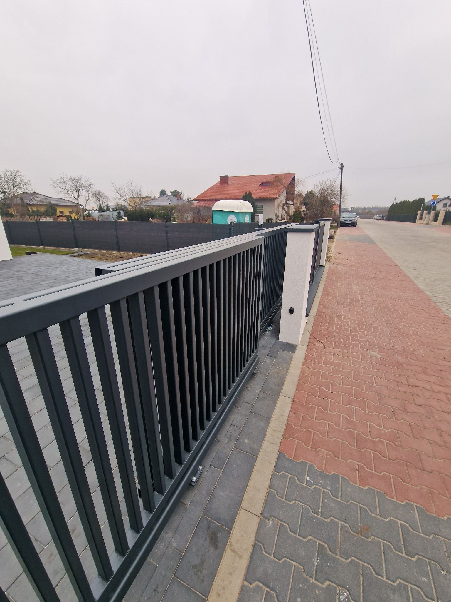 Nowoczesna, przesuwna brama wjazdowa w kolorze antracytowym z pionowymi sztachetkami, zamontowana na posesji z kostką brukową i widokiem na ulicę oraz domy w tle, uchwycona w pochmurny dzień.