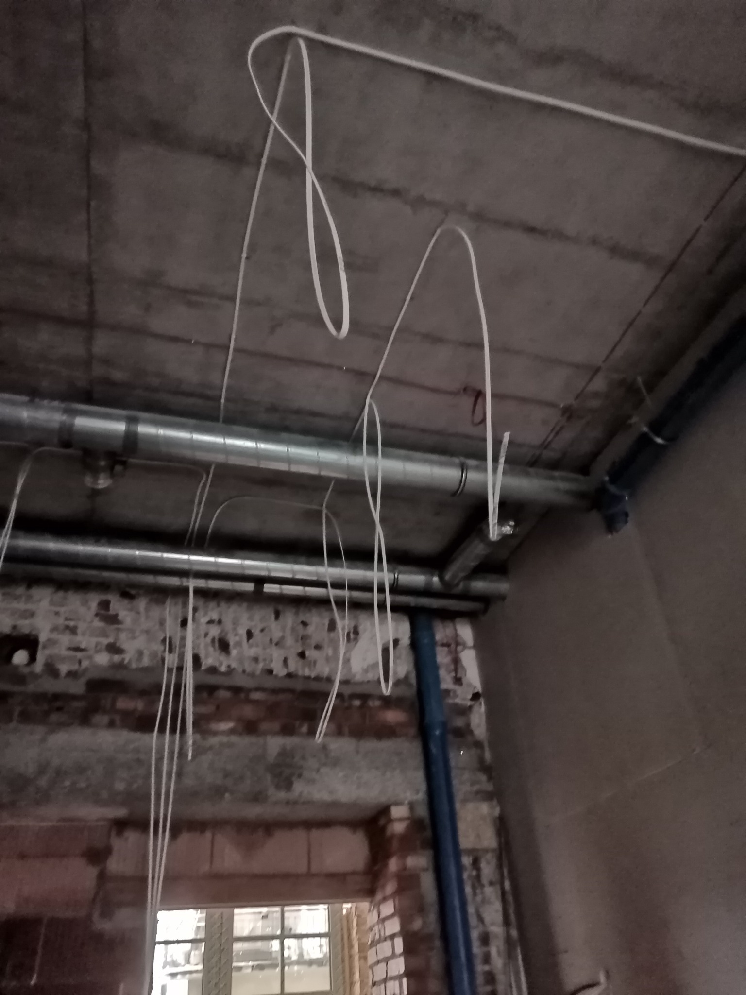 Surowy betonowy sufit z widocznymi rurami wentylacyjnymi i nieułożonymi przewodami elektrycznymi zwisającymi luźno; w tle fragment okna i muru z cegły.