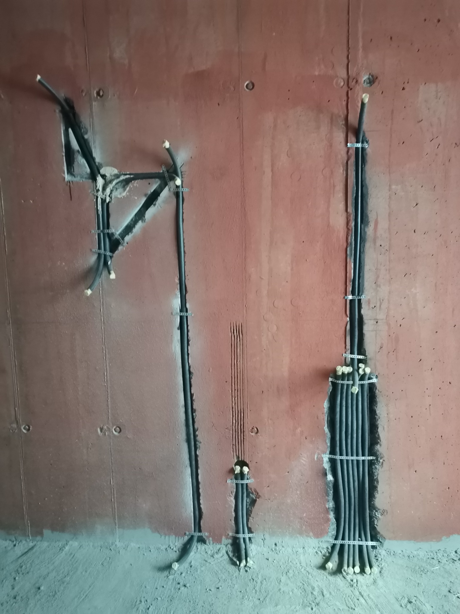 Przewody elektryczne w peszlach zamocowane do surowej, czerwonej ściany w trakcie prac instalacyjnych. Widoczne kable prowadzone w bruzdach, zabezpieczone metalowymi uchwytami. Fragment podłogi...