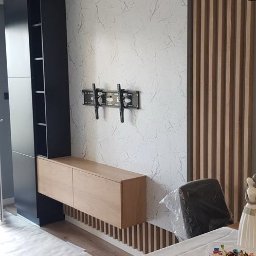 Remonty AZ - Nowoczesna aranżacja ściany z marmurową tapetą, drewnianymi lamelami i szafką RTV. Widoczny uchwyt na telewizor. Podłoga zabezpieczona folią malarską.