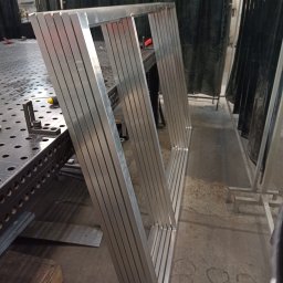DD-db - Aluminiowa konstrukcja ramowa z profili, spawana na stole spawalniczym. Widoczne ślady po spawaniu i zaciski. Tło stanowią zasłony spawalnicze.