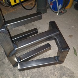 DD-db - Metalowa konstrukcja z profili kwadratowych, spawana, surowa, leżąca na betonowej podłodze. Widoczne spawy i niedoskonałości obróbki. W tle narzędzia i akcesoria warsztatowe.