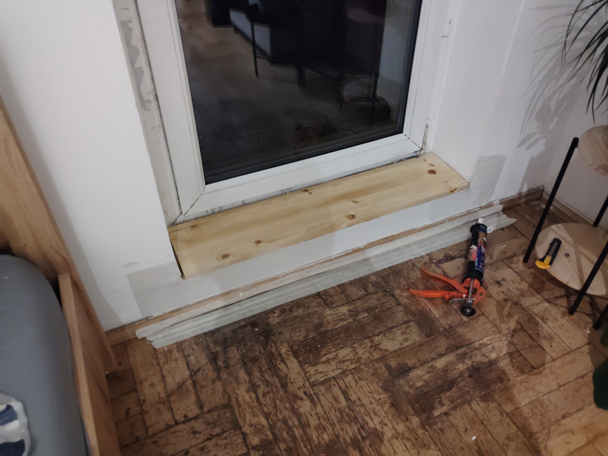 Montaż drewnianego parapetu wewnętrznego przy oknie PCV. Widoczny pistolet do silikonu i listwy przypodłogowe. Parkiet w jodełkę.