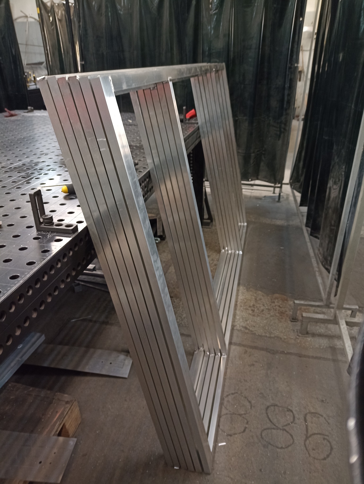 Aluminiowa konstrukcja ramowa z profili, spawana na stole spawalniczym. Widoczne ślady po spawaniu i zaciski. Tło stanowią zasłony spawalnicze.