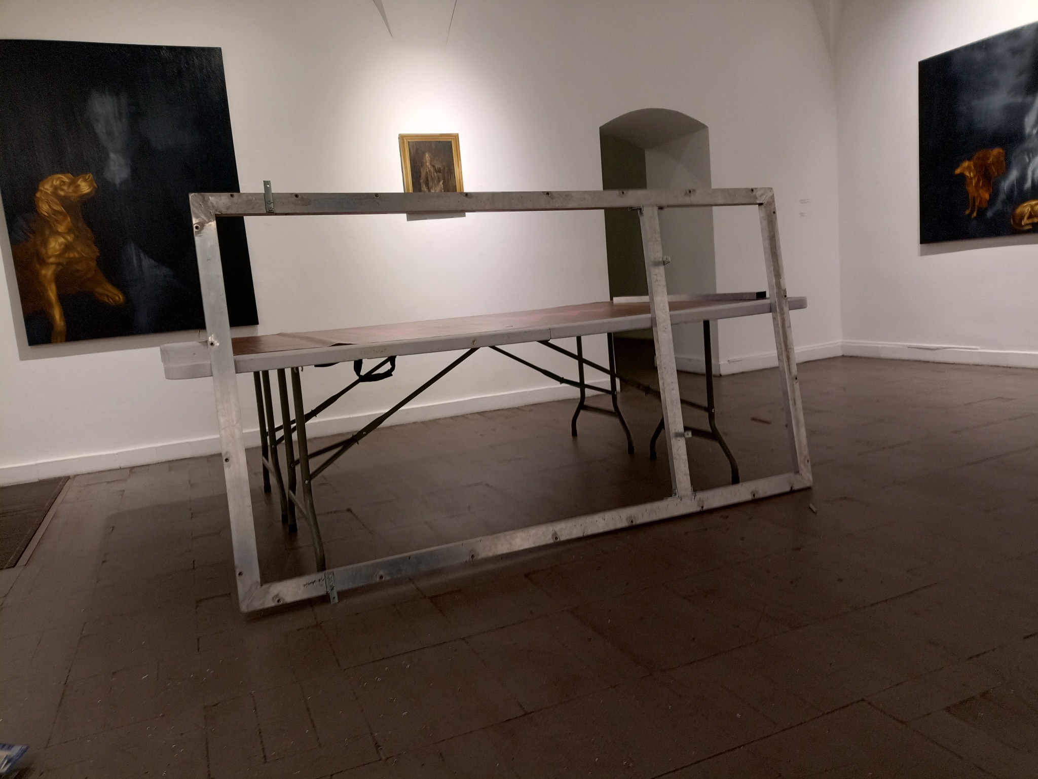 Metalowa konstrukcja ramy na stole w galerii sztuki, z obrazami w tle. Surowe wykończenie metalu kontrastuje z artystycznym otoczeniem. Nietypowe ujęcie.