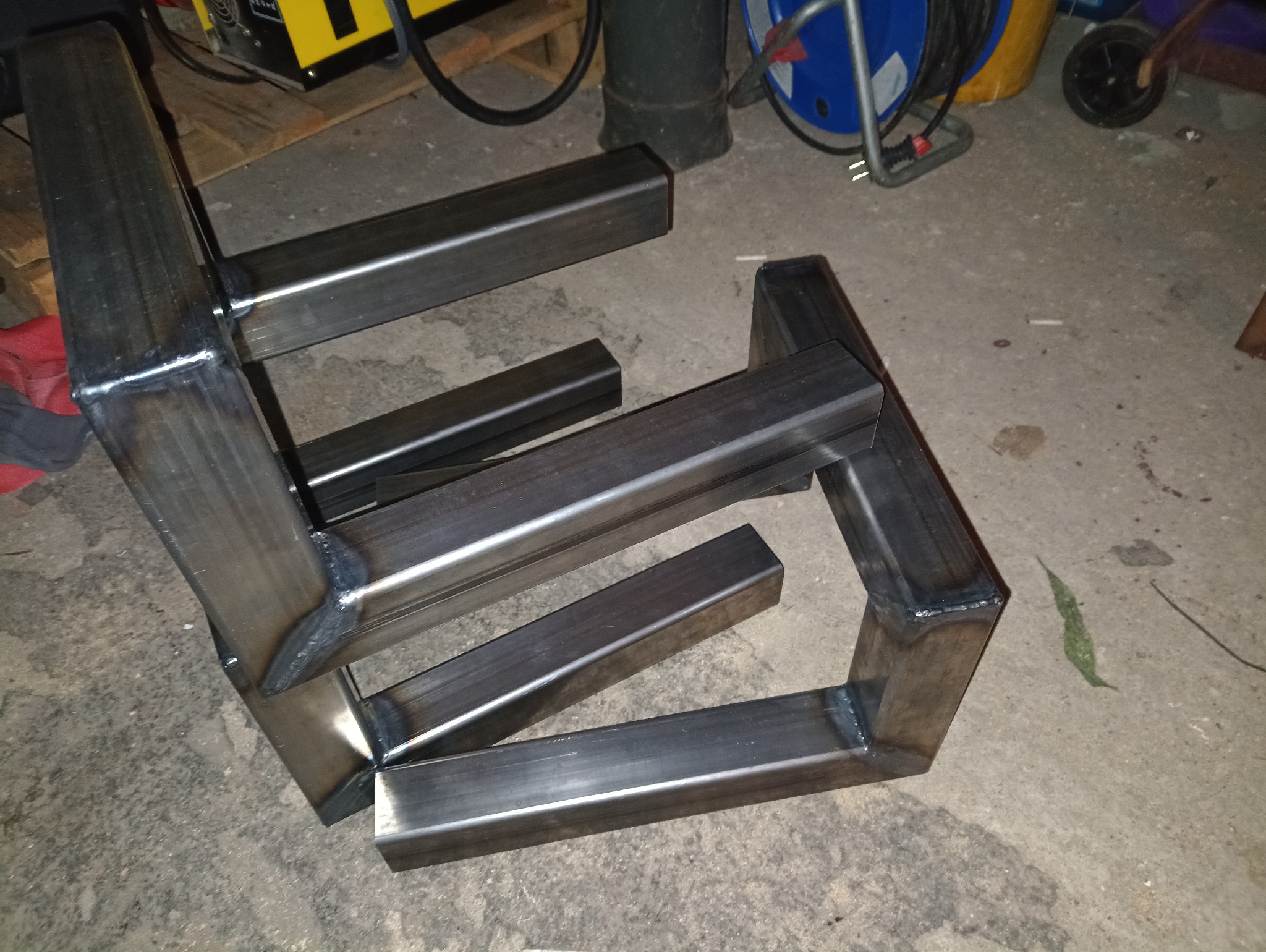 Metalowa konstrukcja z profili kwadratowych, spawana, surowa, leżąca na betonowej podłodze. Widoczne spawy i niedoskonałości obróbki. W tle narzędzia i akcesoria warsztatowe.