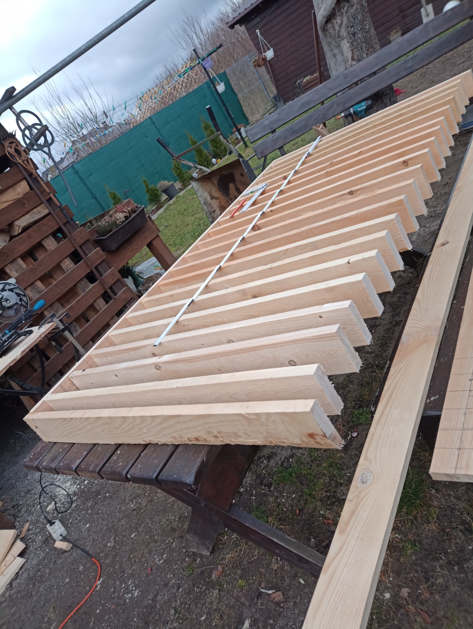 Drewniana konstrukcja z listew na stole, w tle ogród. Elementy konstrukcji ułożone równolegle, tworząc ażurowy wzór. Widoczne narzędzia i materiały przygotowane do dalszej pracy.