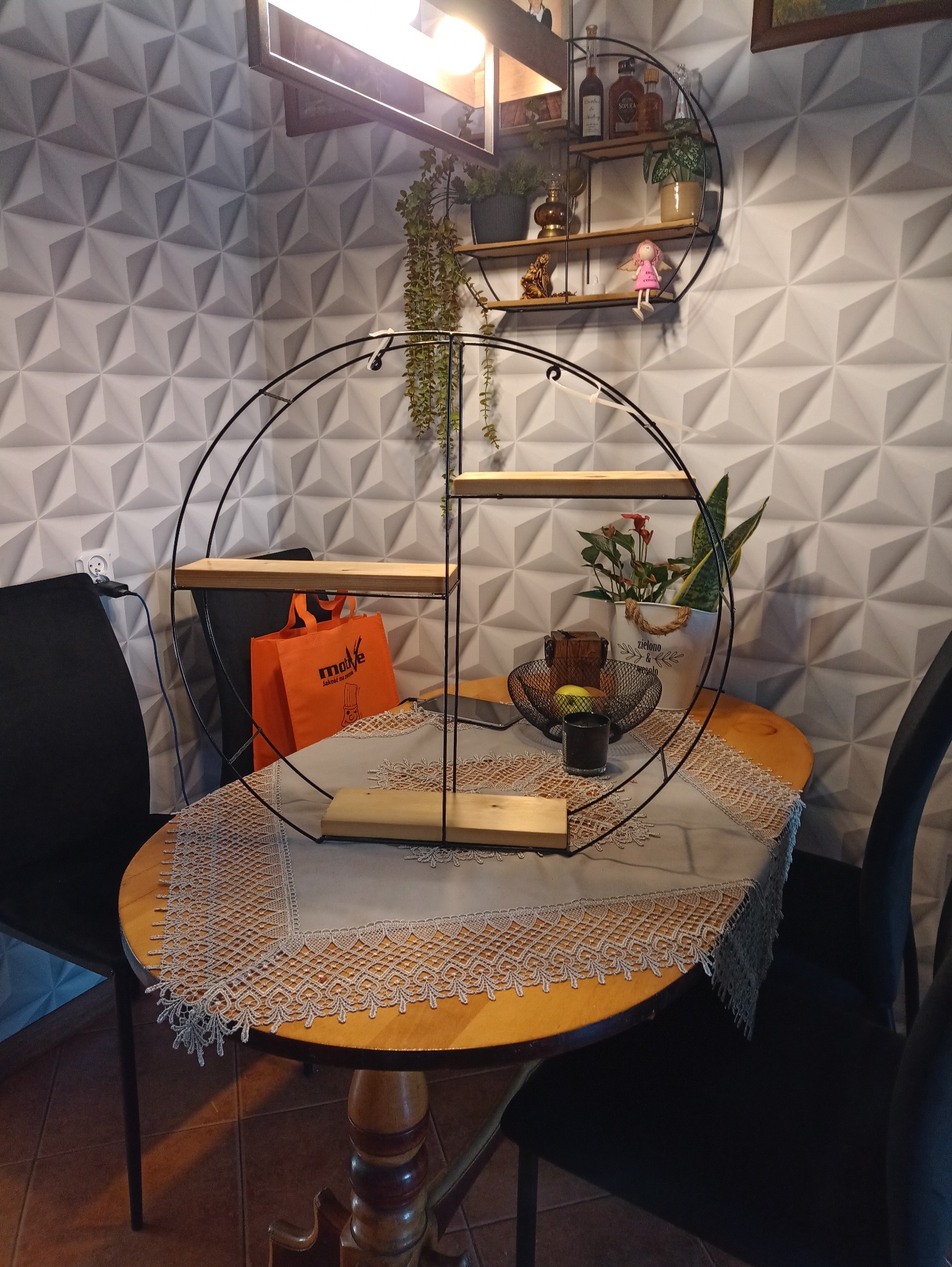 Okrągła, metalowa półka z drewnianymi blatami na stole, dekorowana kwiatami i owocami. W tle trójwymiarowa ściana i wisząca półka z dekoracjami.