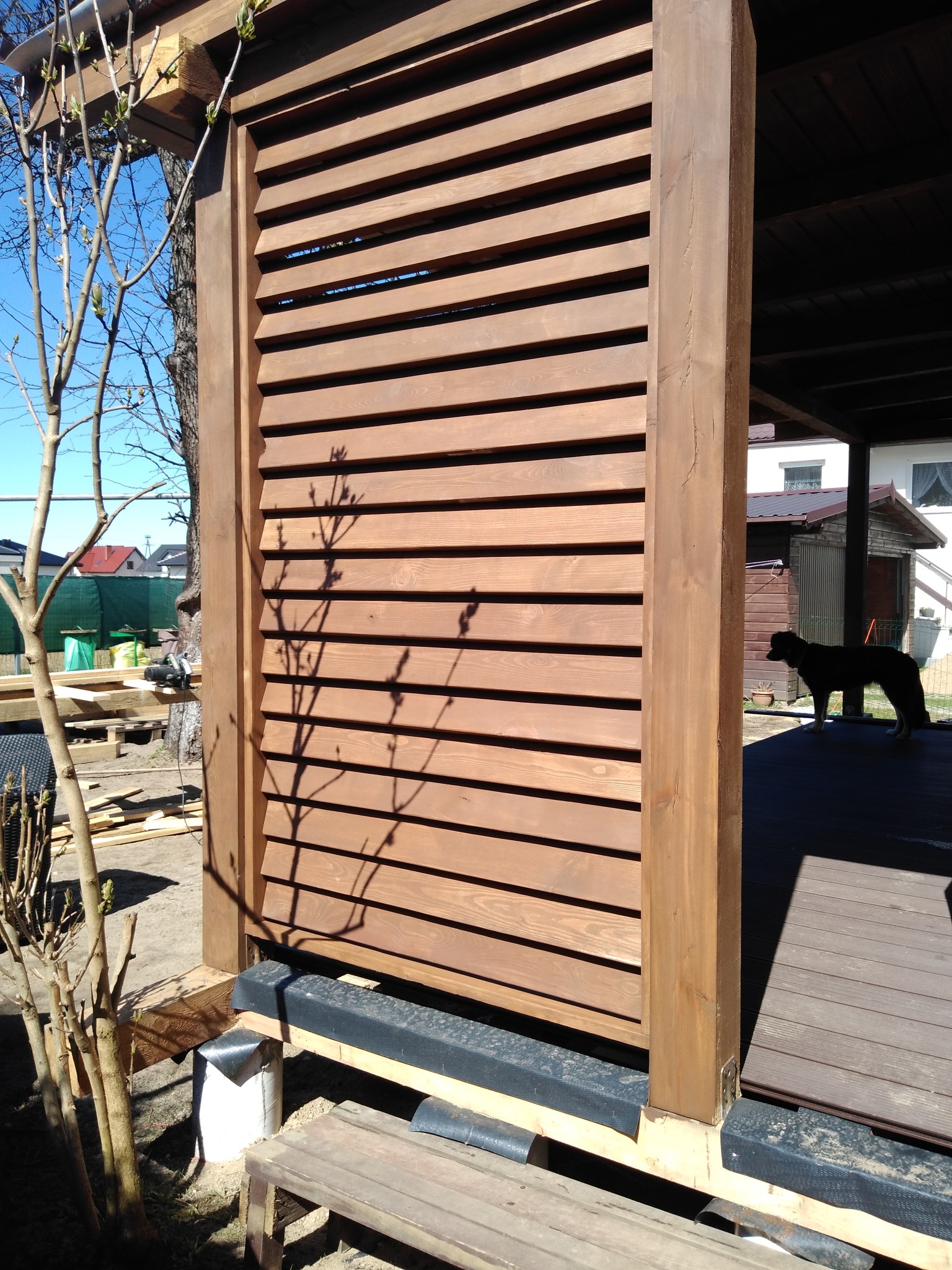 Drewniana konstrukcja z lamelami, prawdopodobnie fragment pergoli lub zabudowy balkonu, z widocznym fragmentem tarasu i psem w tle.