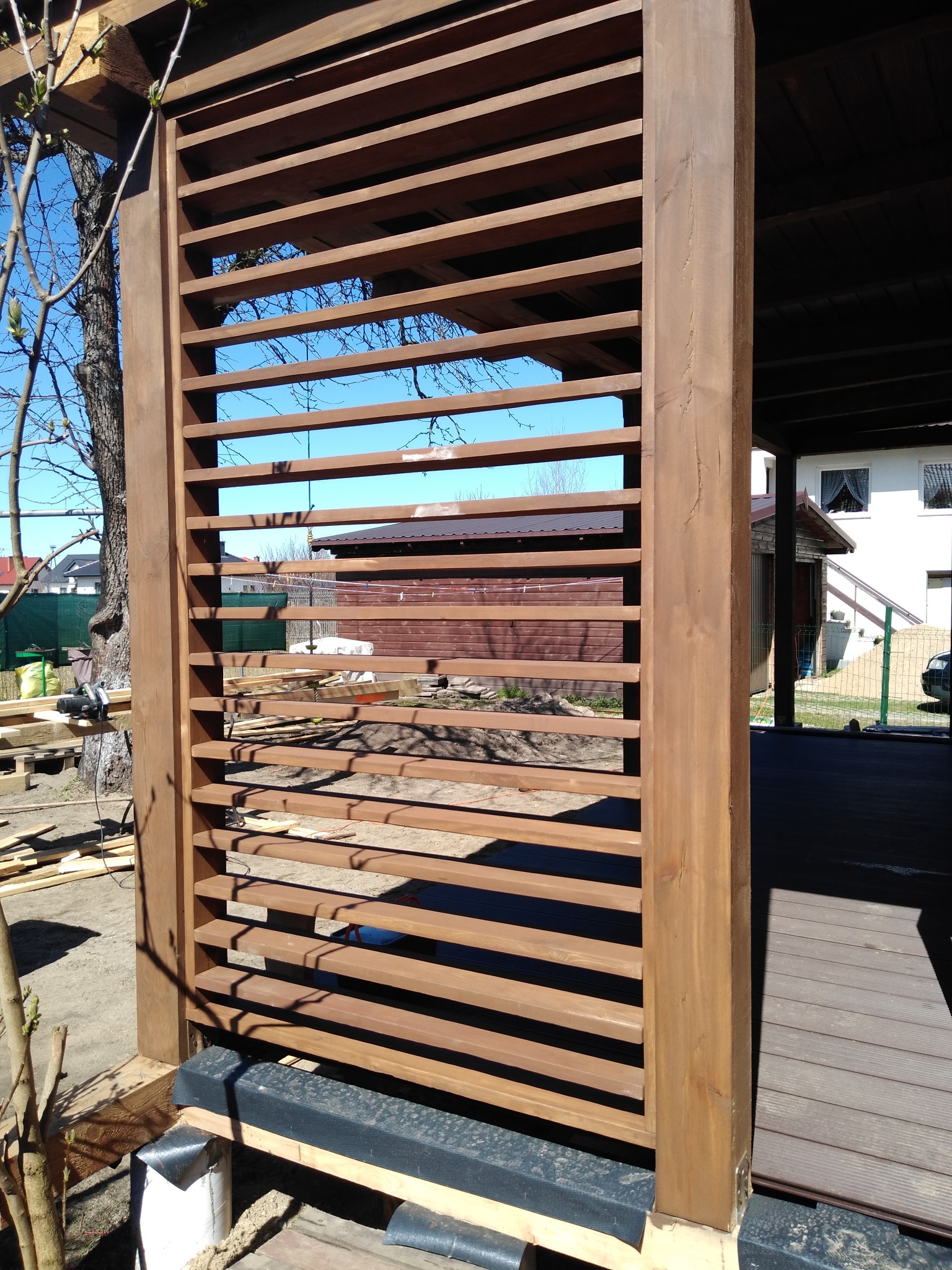 Drewniana konstrukcja z poziomymi listwami, prawdopodobnie fragment pergoli lub tarasu, w trakcie montażu. Widoczne elementy mocujące i narzędzia w tle.