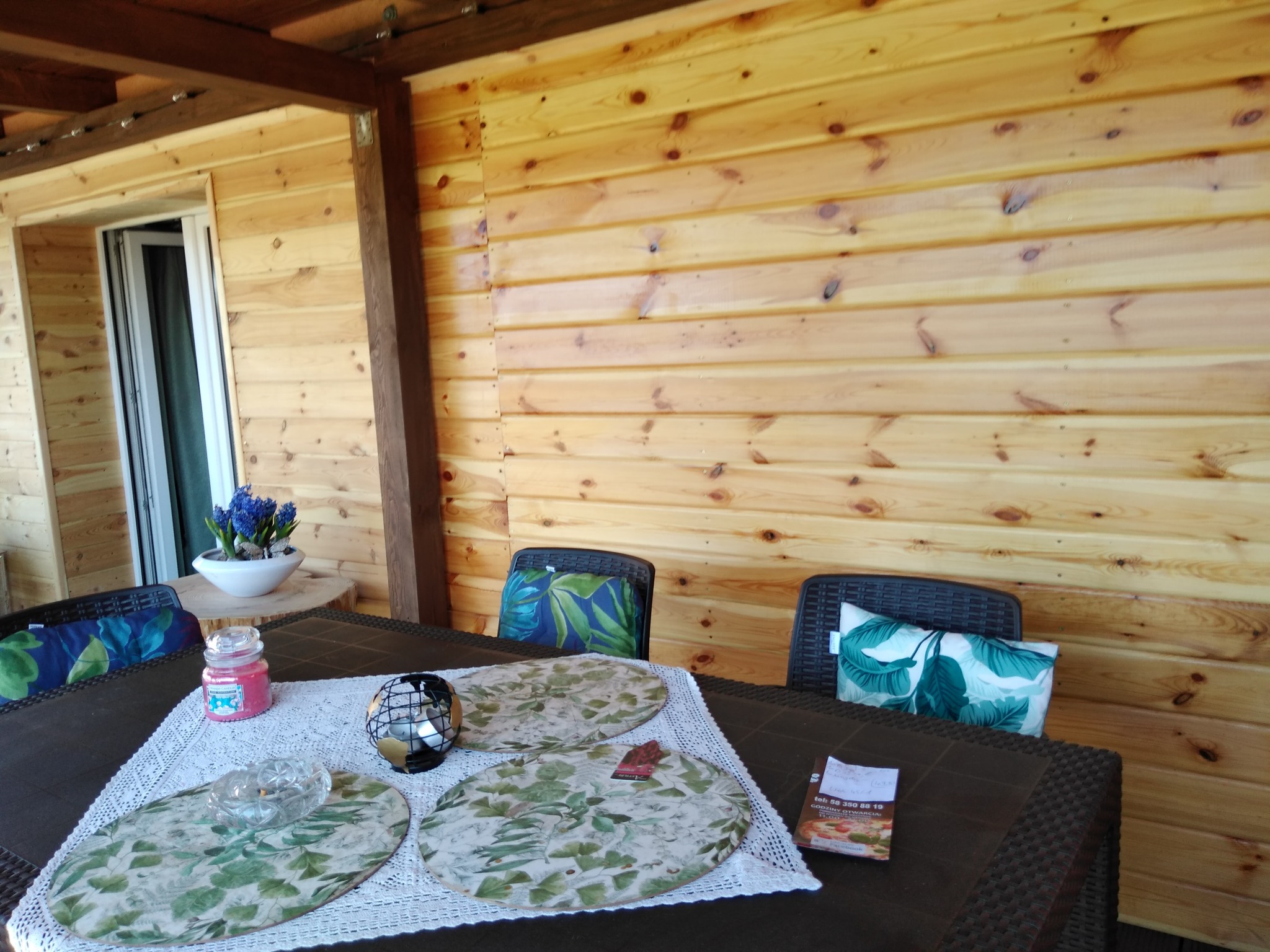 Drewniana zabudowa balkonu z widocznymi deskami boazeryjnymi, stół z dekoracjami i krzesła z poduszkami. Przytulna aranżacja balkonu.