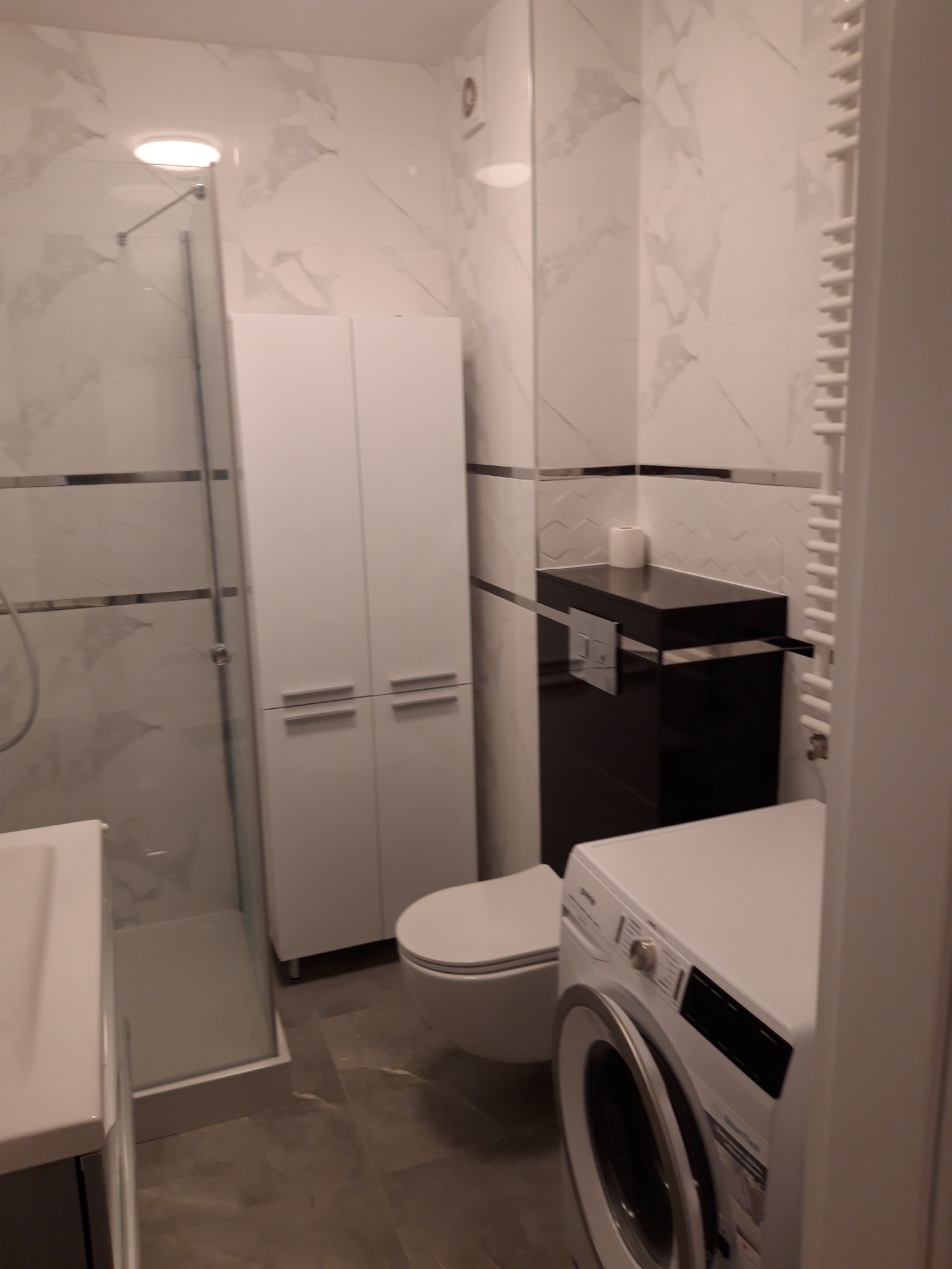 Nowoczesna łazienka z kabiną prysznicową, białą szafką, toaletą, czarną szafką pod umywalkę i pralką, wykończona marmurowymi płytkami z szarymi żyłami.