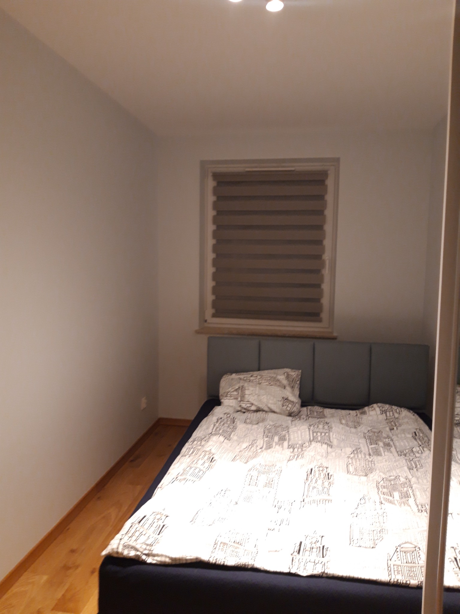 Sypialnia w Warszawie z łóżkiem z szarym wezgłowiem i pościelą w budynki, widok na okno z żaluzjami poziomymi.