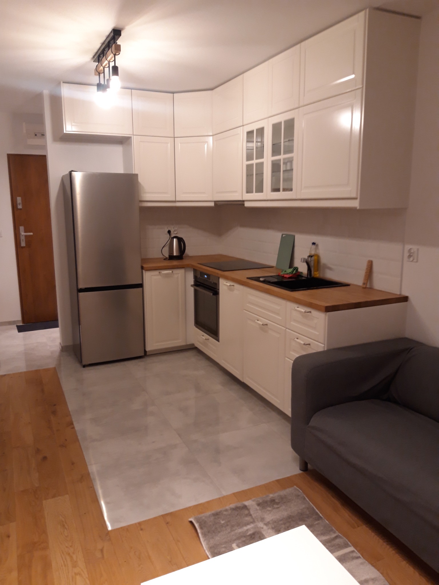 Jasna kuchnia w mieszkaniu z drewnianym blatem, białymi szafkami i szarą sofą w tle. Widoczny nowoczesny plafon z żarówkami.