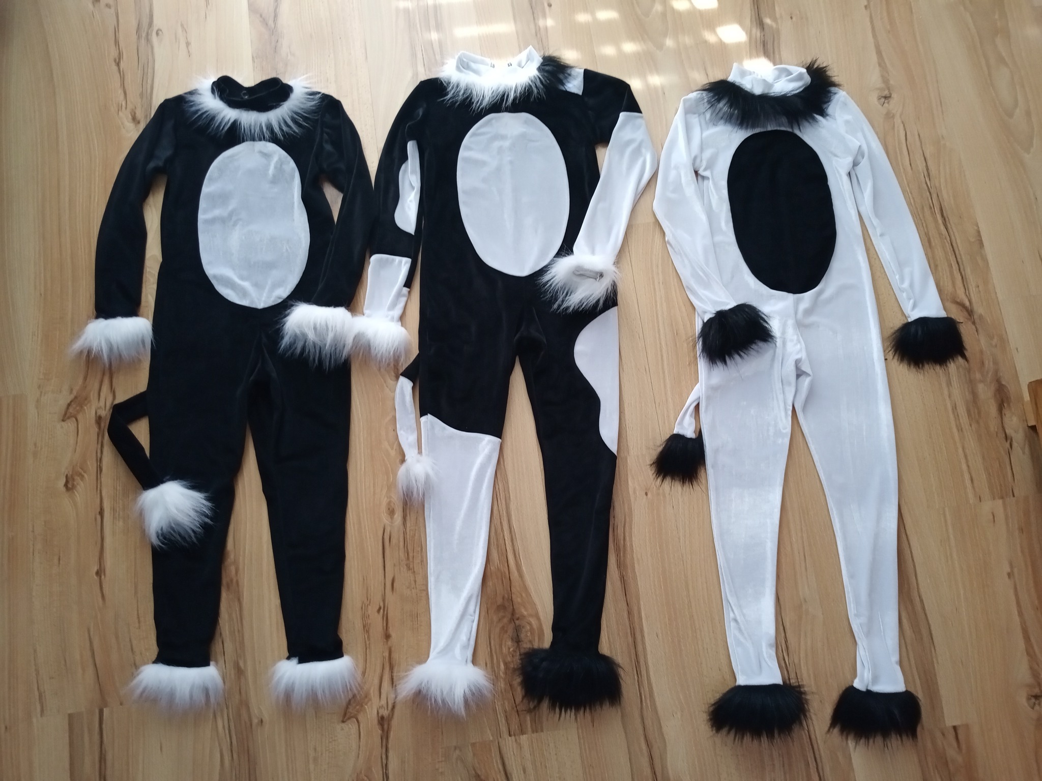 Trzy kostiumy zwierząt w kolorach czerni, bieli i szarości, leżące na drewnianej podłodze, wykończone futerkiem przy szyi, mankietach i nogawkach, z ogonami.