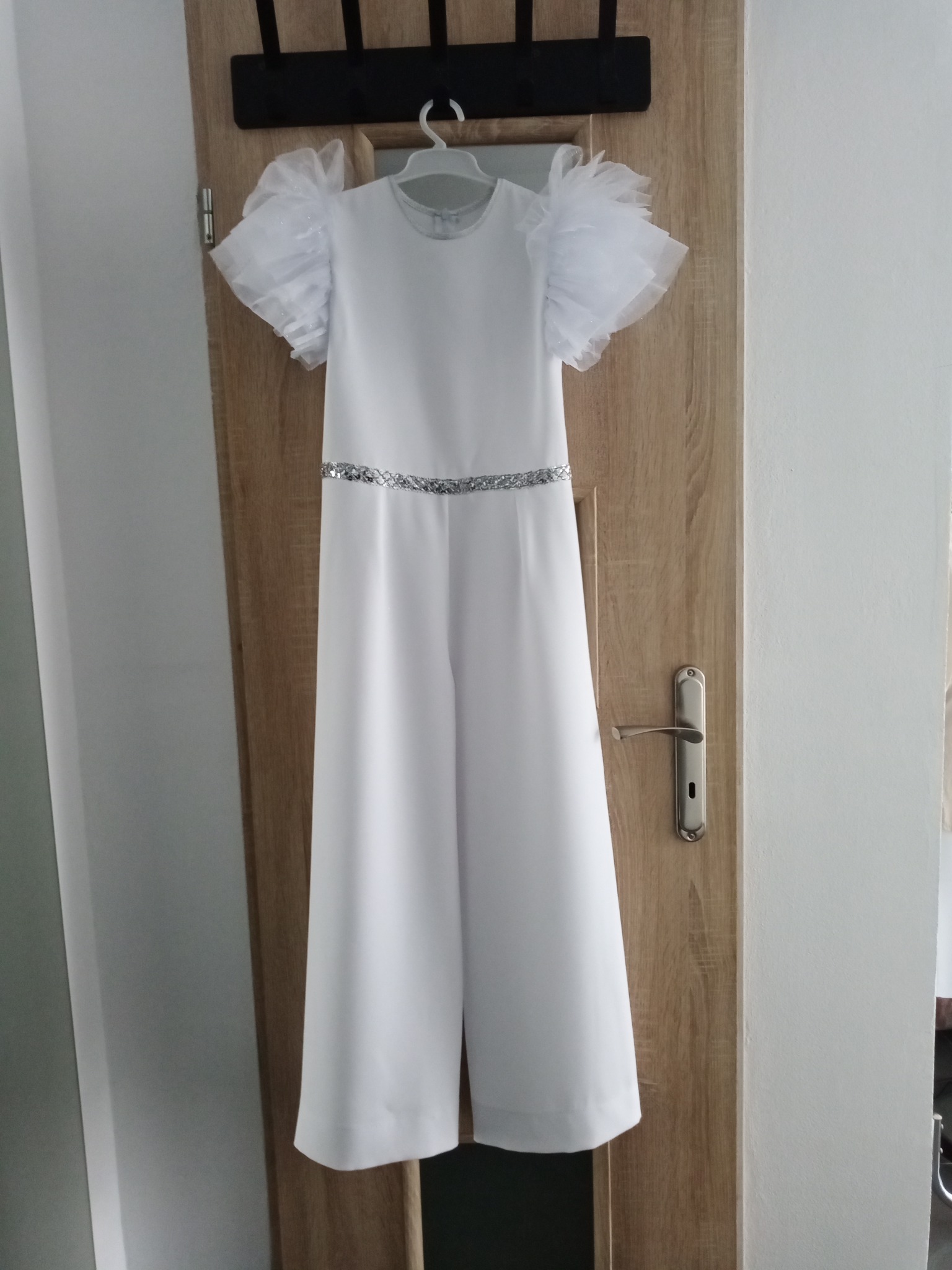 Elegancki biały kombinezon damski z krótkimi rękawami z tiulowymi bufkami i srebrnym paskiem, wiszący na wieszaku na drzwiach.