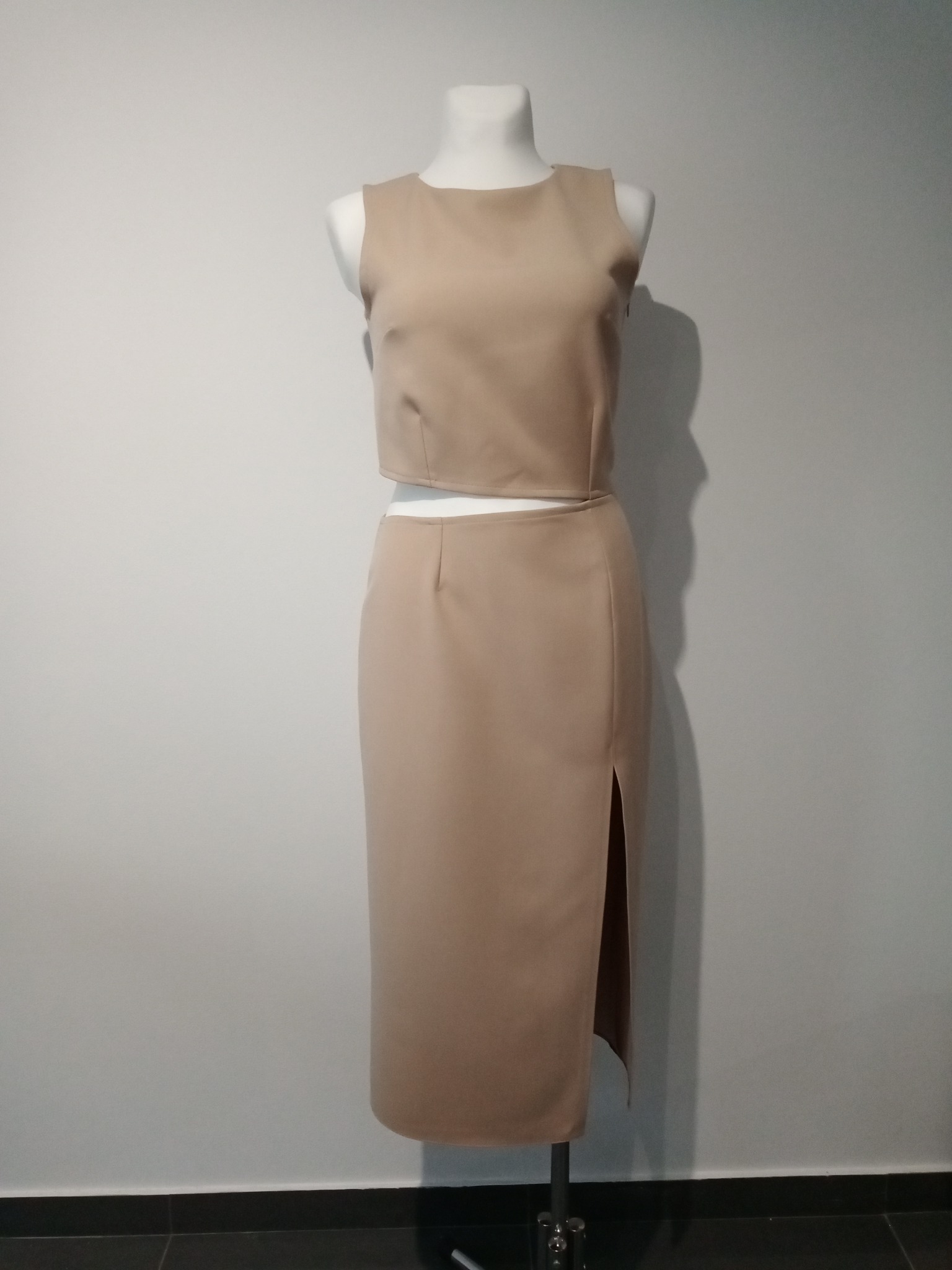 Beżowy komplet damski: krótki top i spódnica midi z rozcięciem, prezentowany na manekinie na tle szarej ściany.