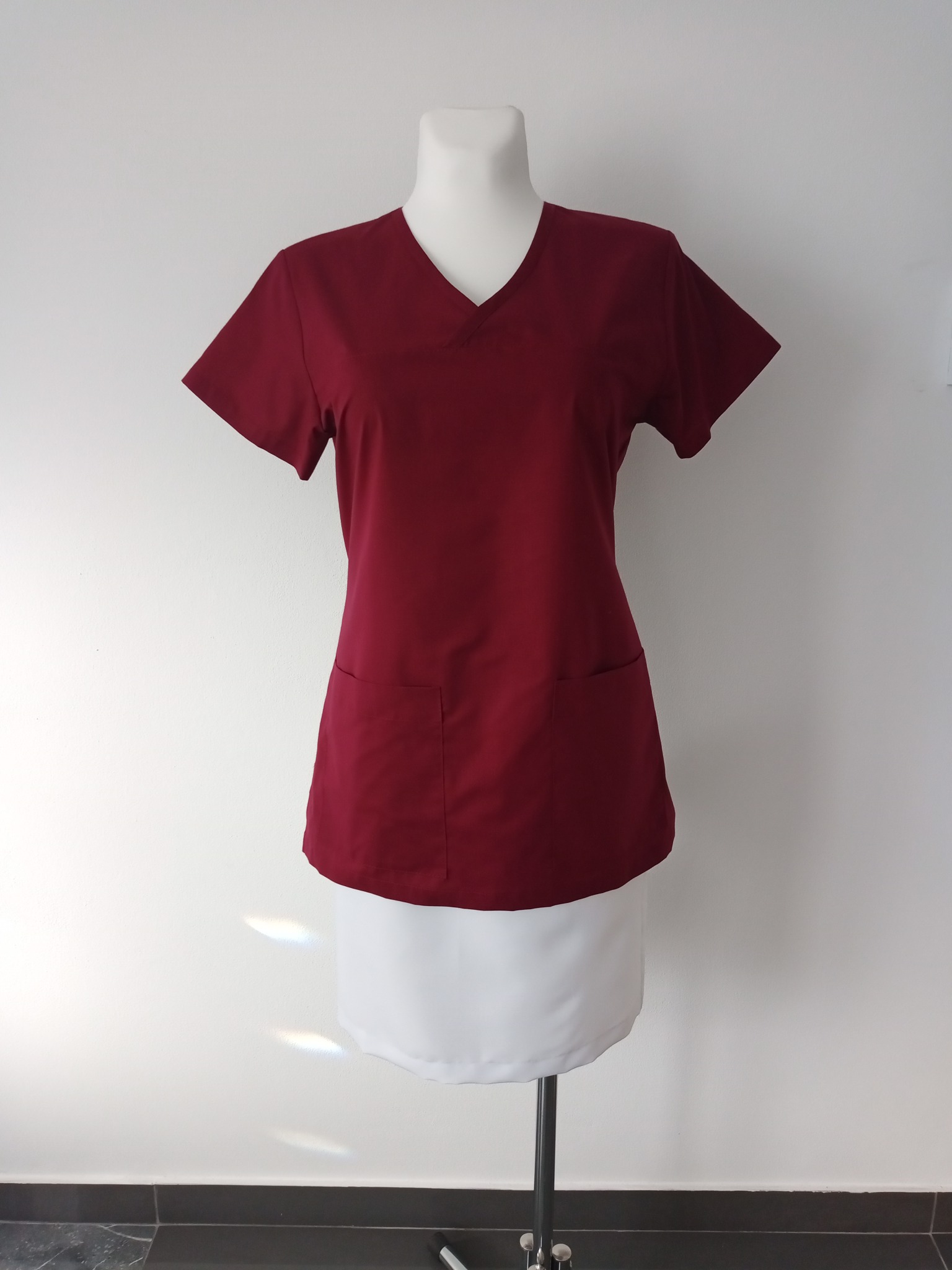 Bordowa bluza medyczna z krótkim rękawem i dwiema kieszeniami, zaprezentowana na manekinie na tle białej ściany i ciemnej podłogi.