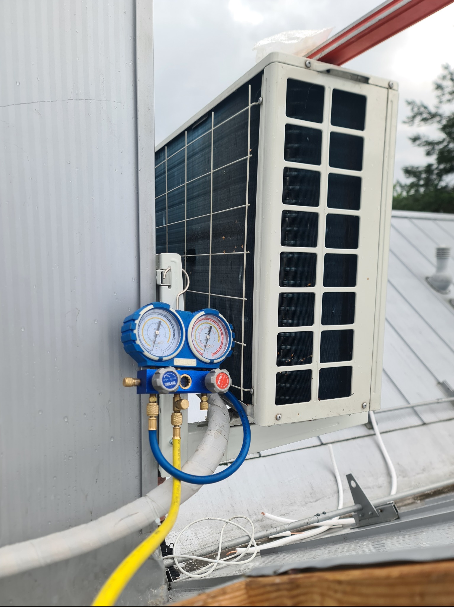 Biała jednostka zewnętrzna klimatyzacji na szarym dachu, podłączona do niebieskiego manometru z żółtymi i niebieskimi przewodami, sugerująca serwis lub instalację.