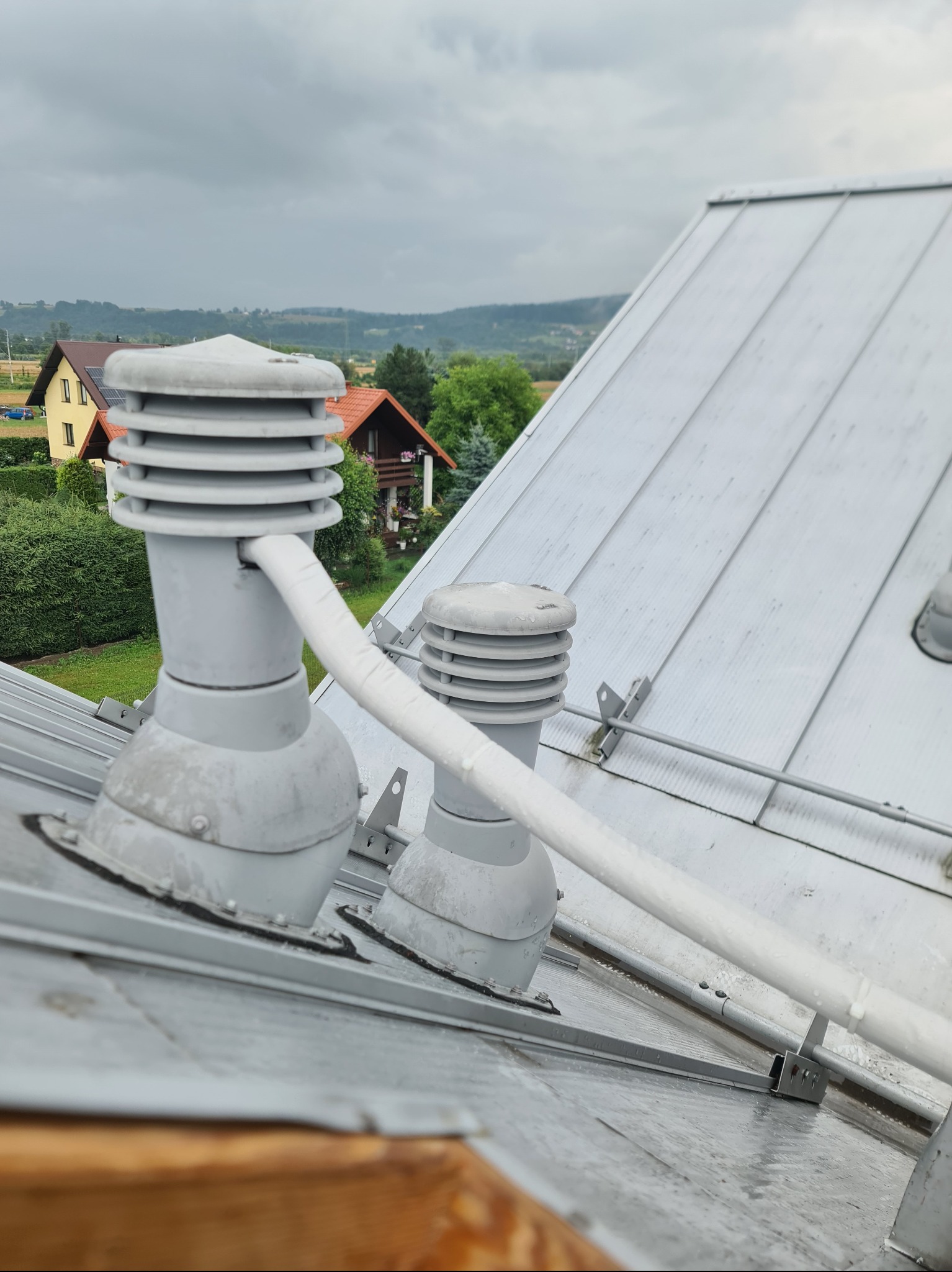 Szare kominki wentylacyjne na metalowym dachu domu jednorodzinnego, z widokiem na okoliczne budynki i pochmurne niebo w tle. Elementy instalacji wentylacyjnej.