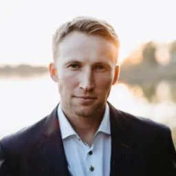 Portret mężczyzny w garniturze na tle jeziora o zachodzie słońca, z delikatnym uśmiechem.