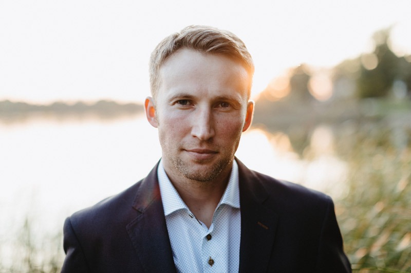 Portret mężczyzny w garniturze na tle jeziora o zachodzie słońca, z delikatnym uśmiechem.