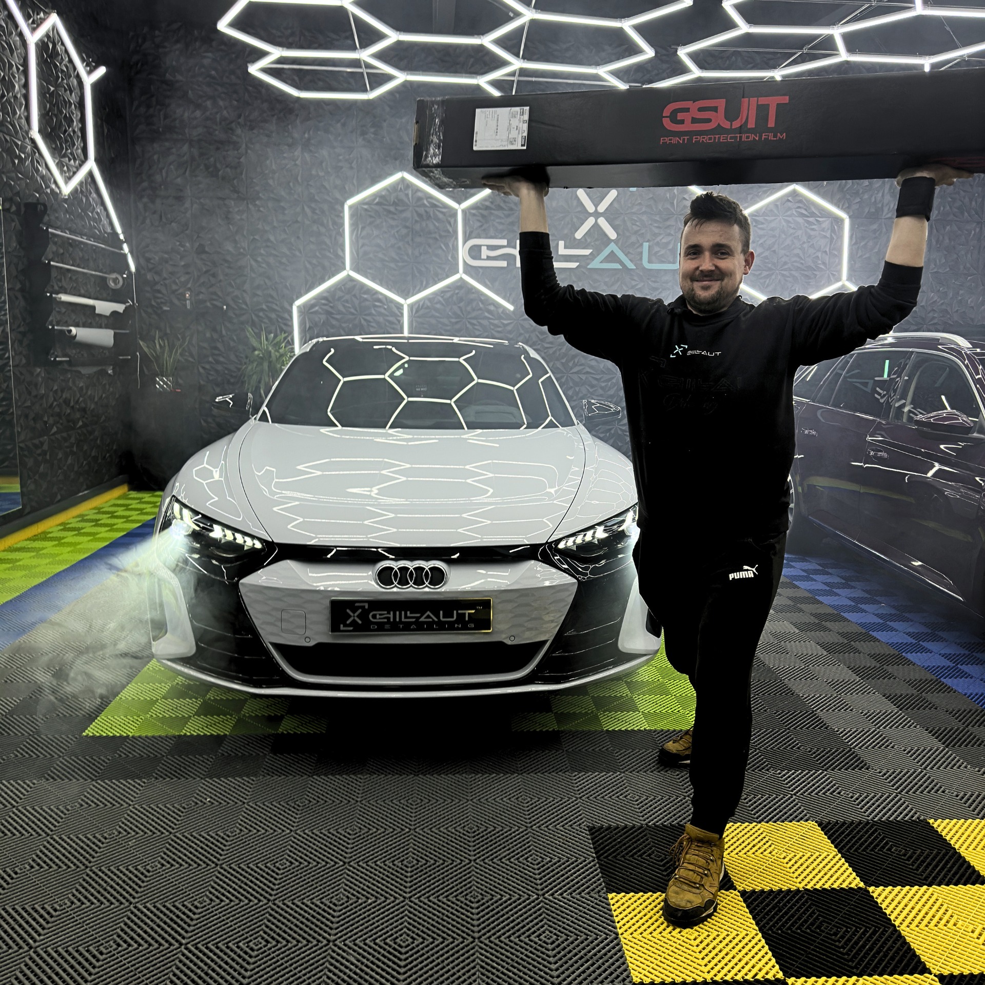 Pracownik firmy z Kościana prezentuje opakowanie folii ochronnej GSuit nad białym Audi e-tron GT w nowoczesnym, oświetlonym studio detailingowym z heksagonalnymi lampami.