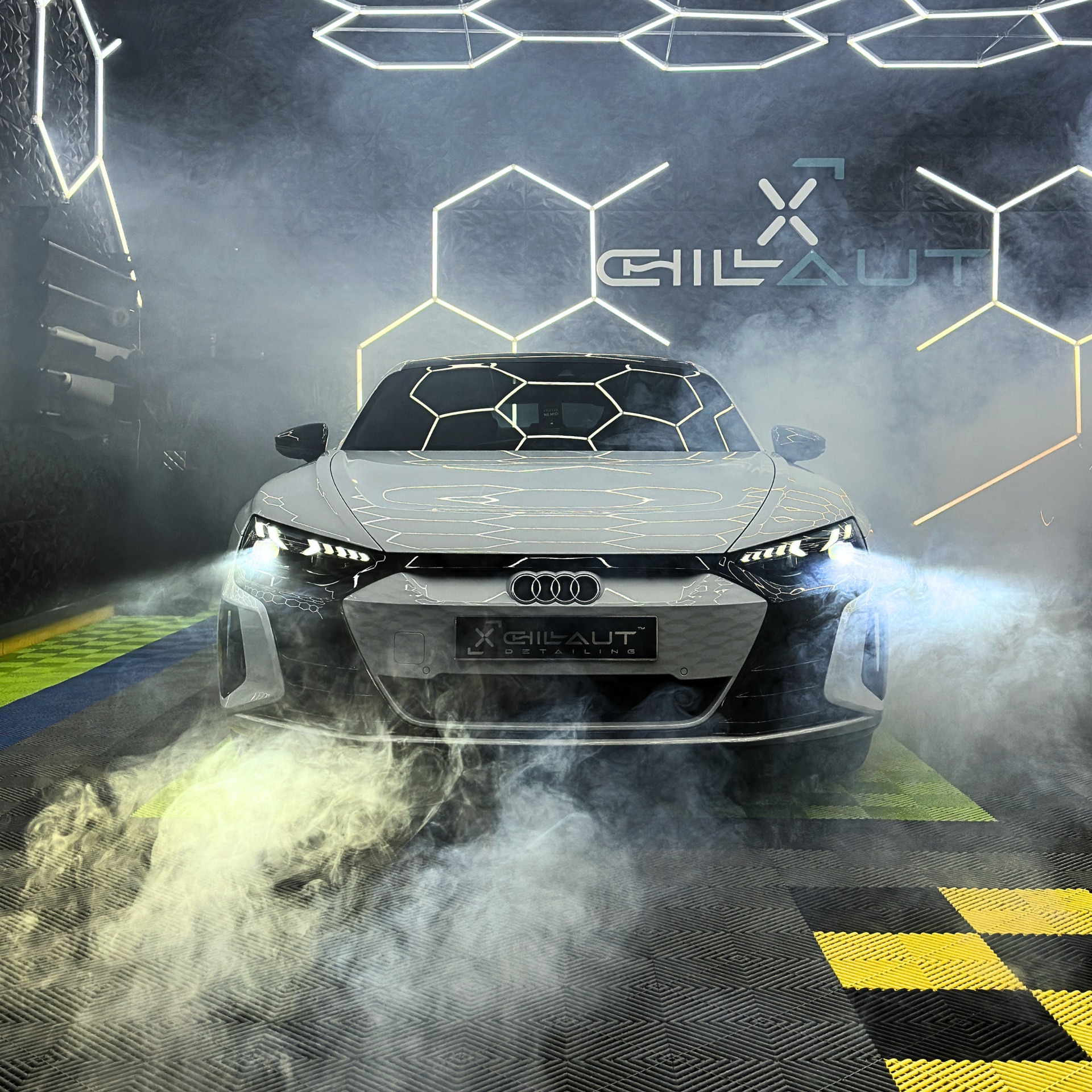 Białe Audi e-tron GT w studiu detailingowym, oświetlone neonowymi heksagonami i otoczone parą, prezentowane od przodu.