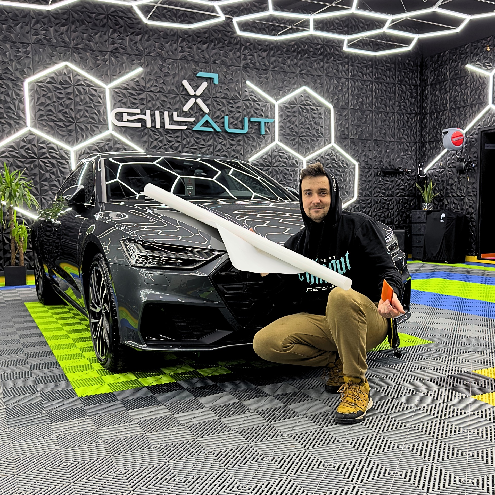 Mężczyzna w bluzie z kapturem prezentuje rolkę folii do oklejania samochodu Audi A7 w nowoczesnym studio detailingowym z heksagonalnymi panelami na ścianie i podświetleniem LED.