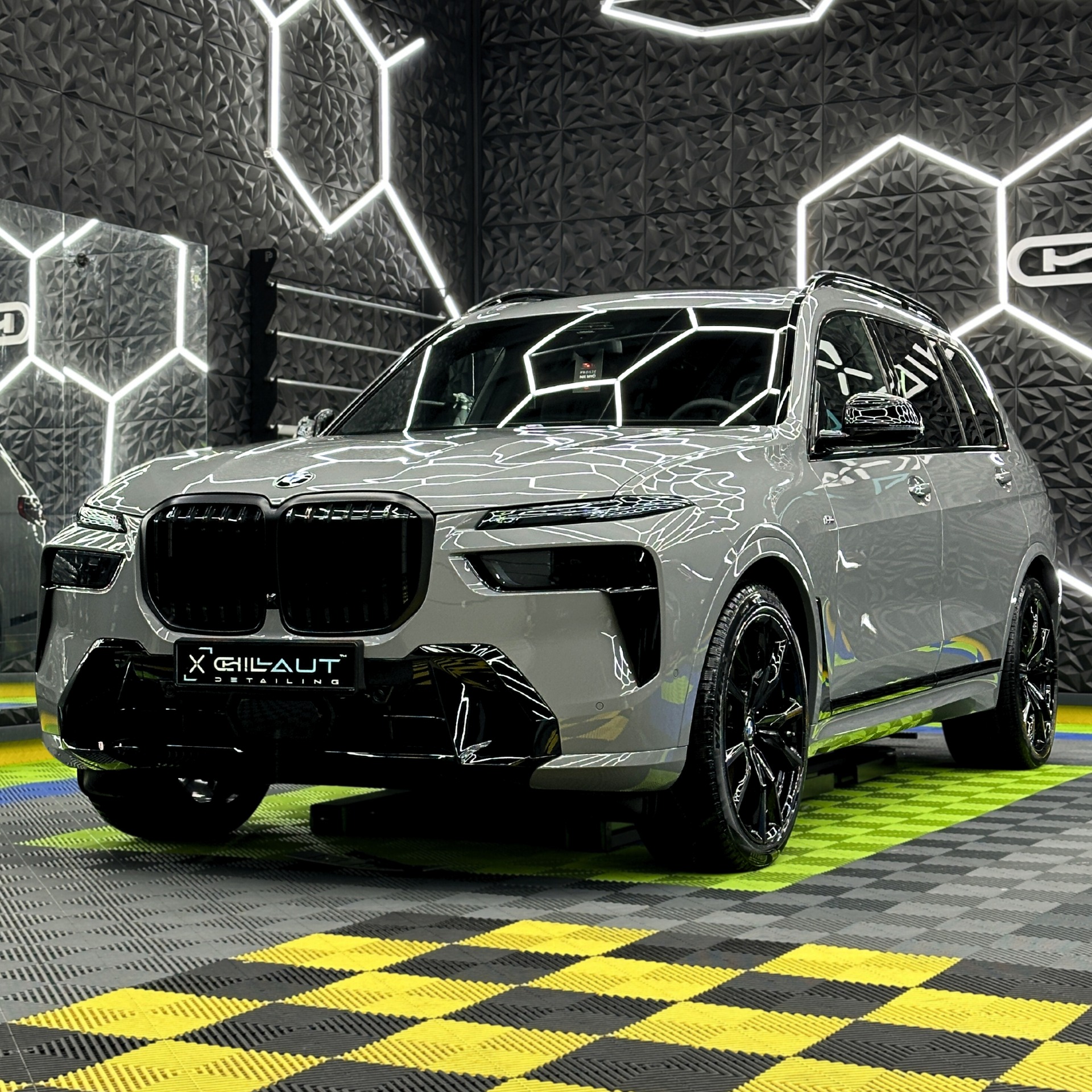 Szary SUV BMW po detailingu, z czarnymi felgami, na podłodze w szachownicę, w nowoczesnym studiu z oświetleniem LED w kształcie heksagonów na ścianie.