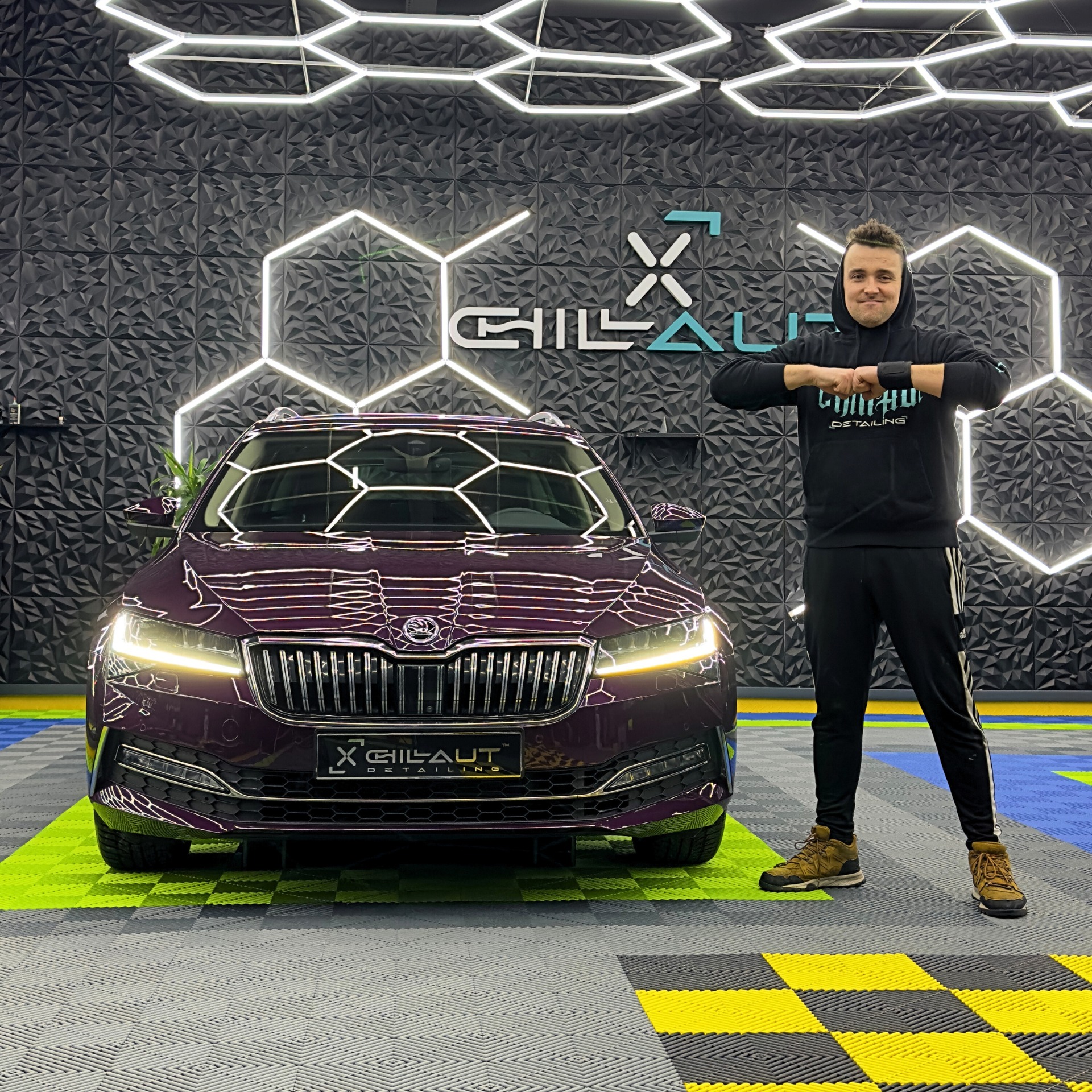 Bordowy samochód Skoda Superb po detailingu, prezentowany przez mężczyznę w bluzie z kapturem z logo firmy na tle ściany z geometrycznym wzorem i oświetleniem LED w kształcie heksagonów.