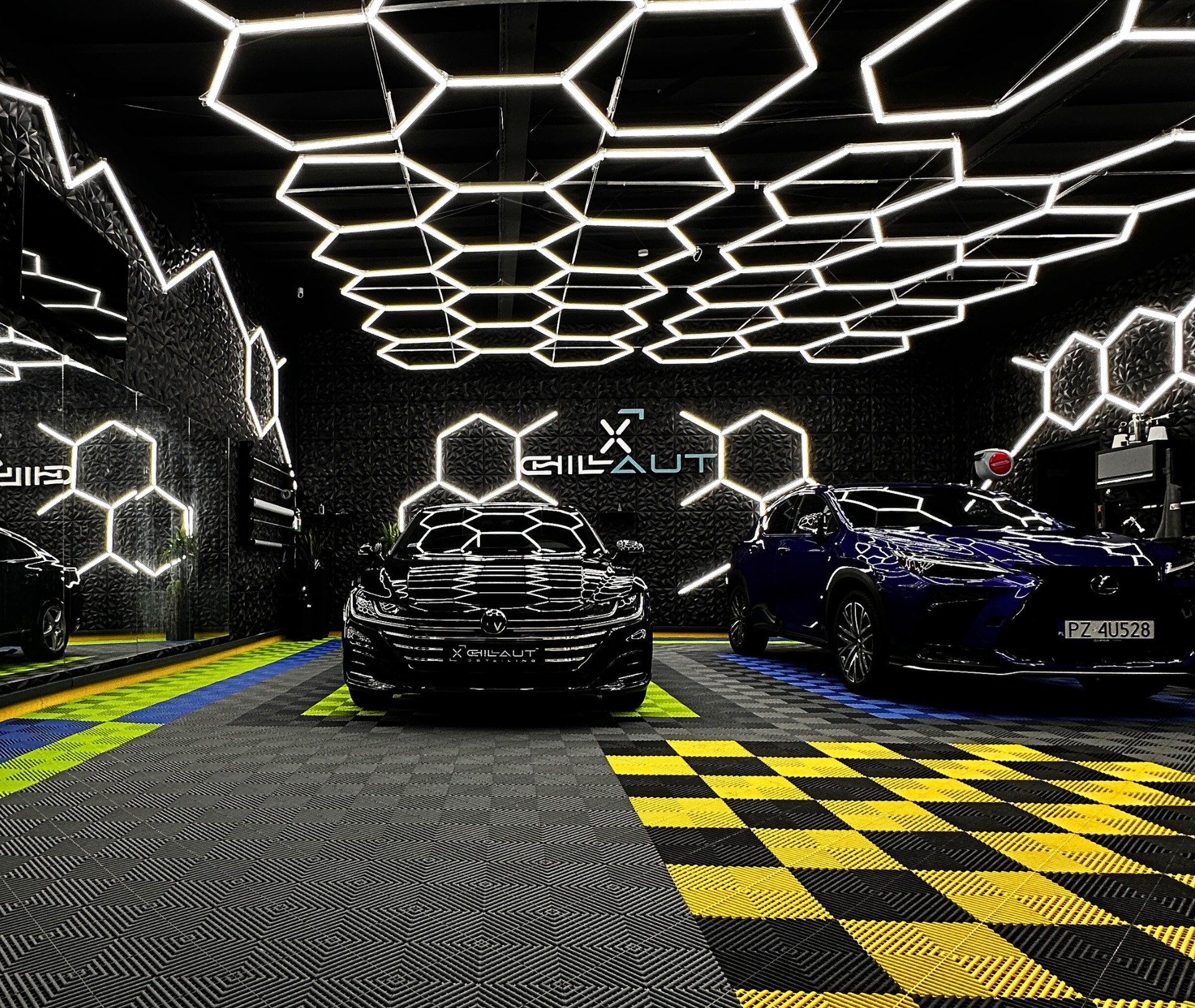 Dwa samochody, czarny Volkswagen i niebieski Lexus, zaparkowane w luksusowym garażu z heksagonalnym oświetleniem LED na suficie i ścianach, z podłogą w szachownicę w kolorach żółtym, czarnym...