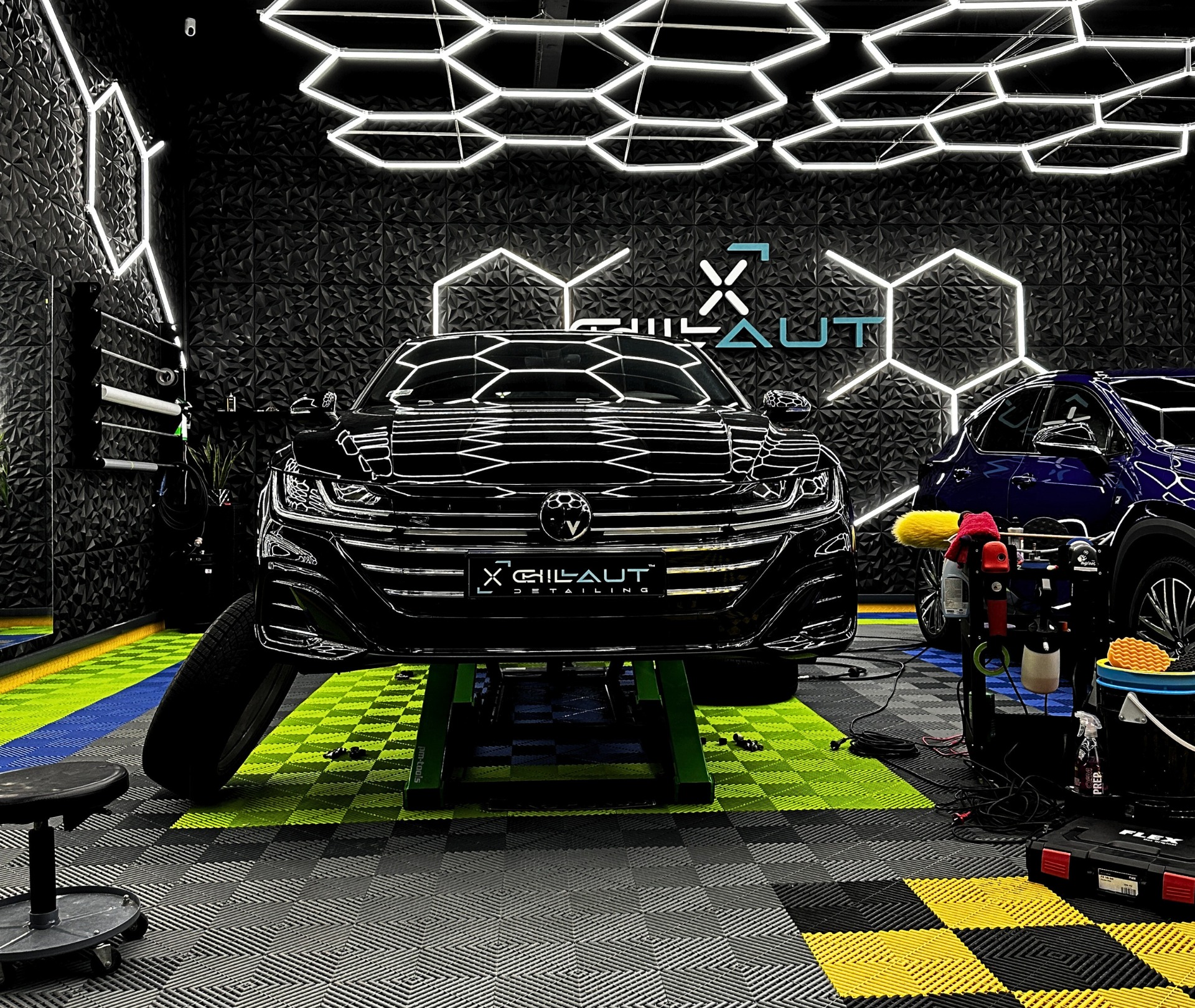 Czarny samochód na podnośniku w studiu detailingowym z heksagonalnym oświetleniem LED i teksturowaną ścianą, widoczne narzędzia i akcesoria do polerowania.