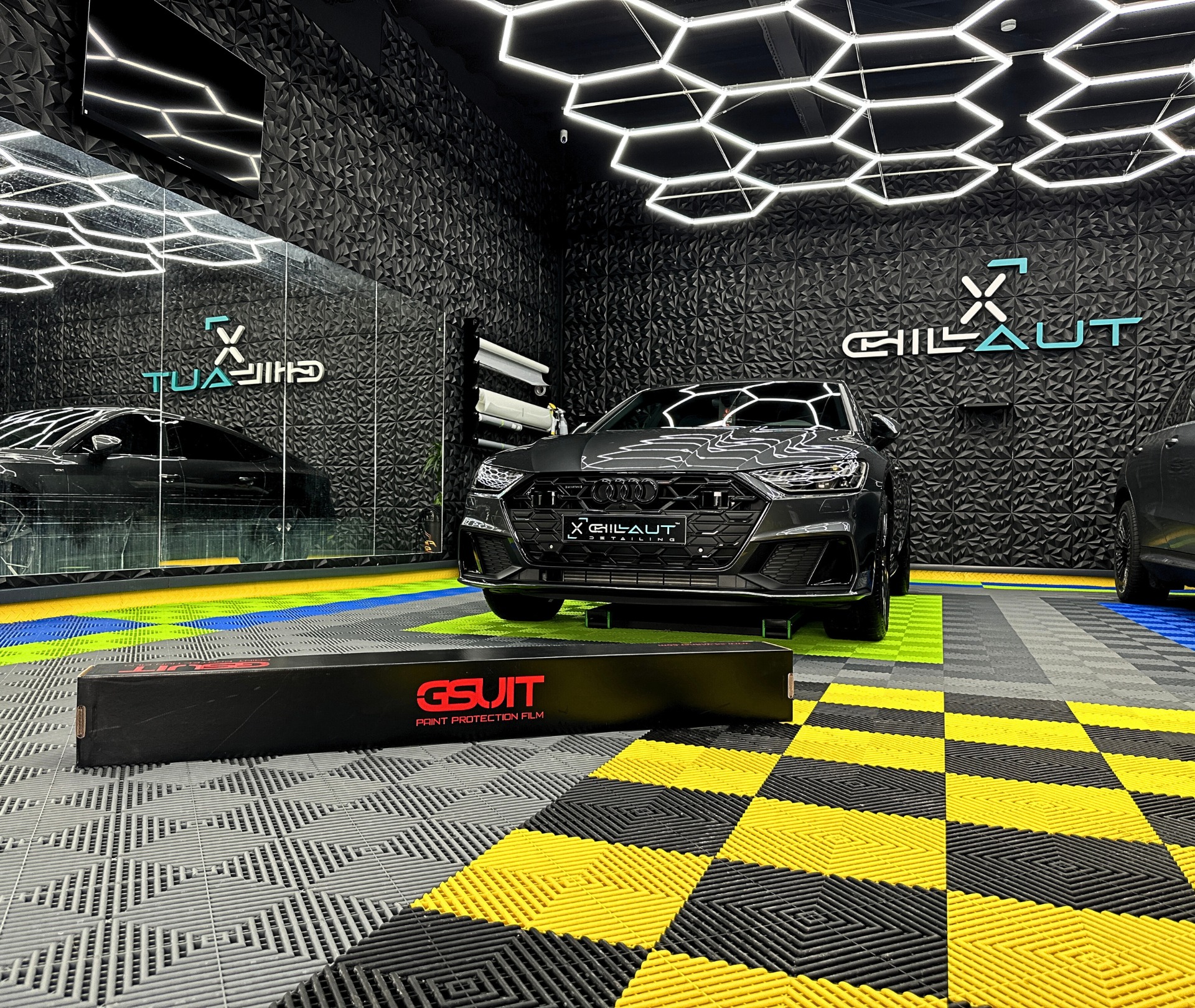 Szary Audi A7 po detailingu w studio z dekoracyjną ścianą 3D, podświetlanym sufitem i podłogą z kolorowych płytek, z widocznym opakowaniem folii ochronnej GSUIT