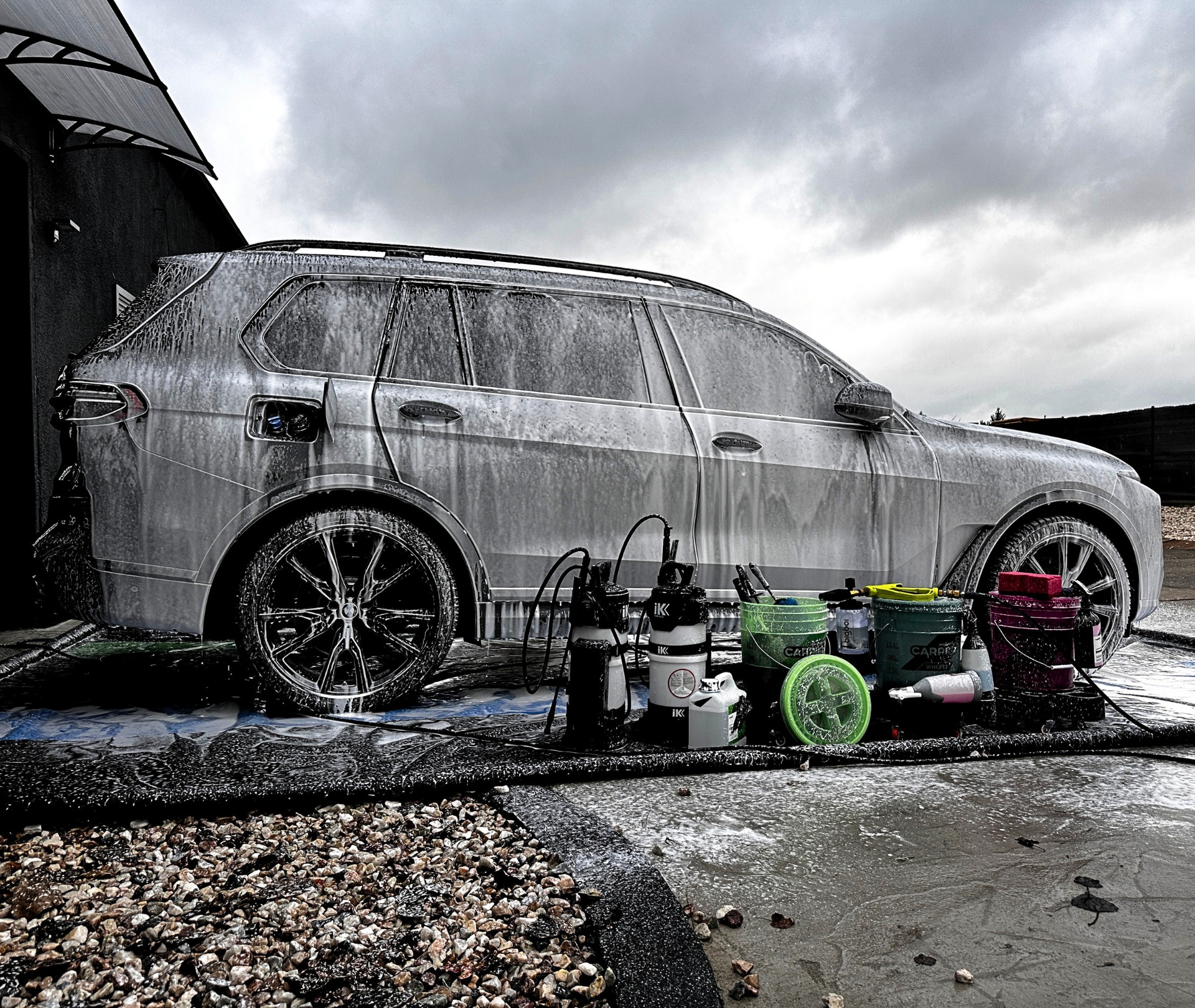 Szary SUV pokryty pianą myjącą, widoczne czarne felgi, obok stoją wiadra, butelki i spryskiwacze do czyszczenia samochodów, w tle pochmurne niebo.