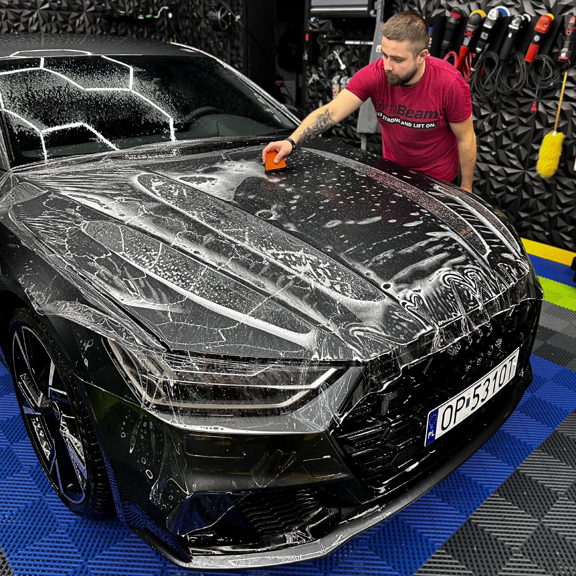 Nakładanie folii ochronnej na czarny samochód Audi A7 w profesjonalnym studiu detailingowym; widoczna ręka detailera z tatuażem i pomarańczowy aplikator.