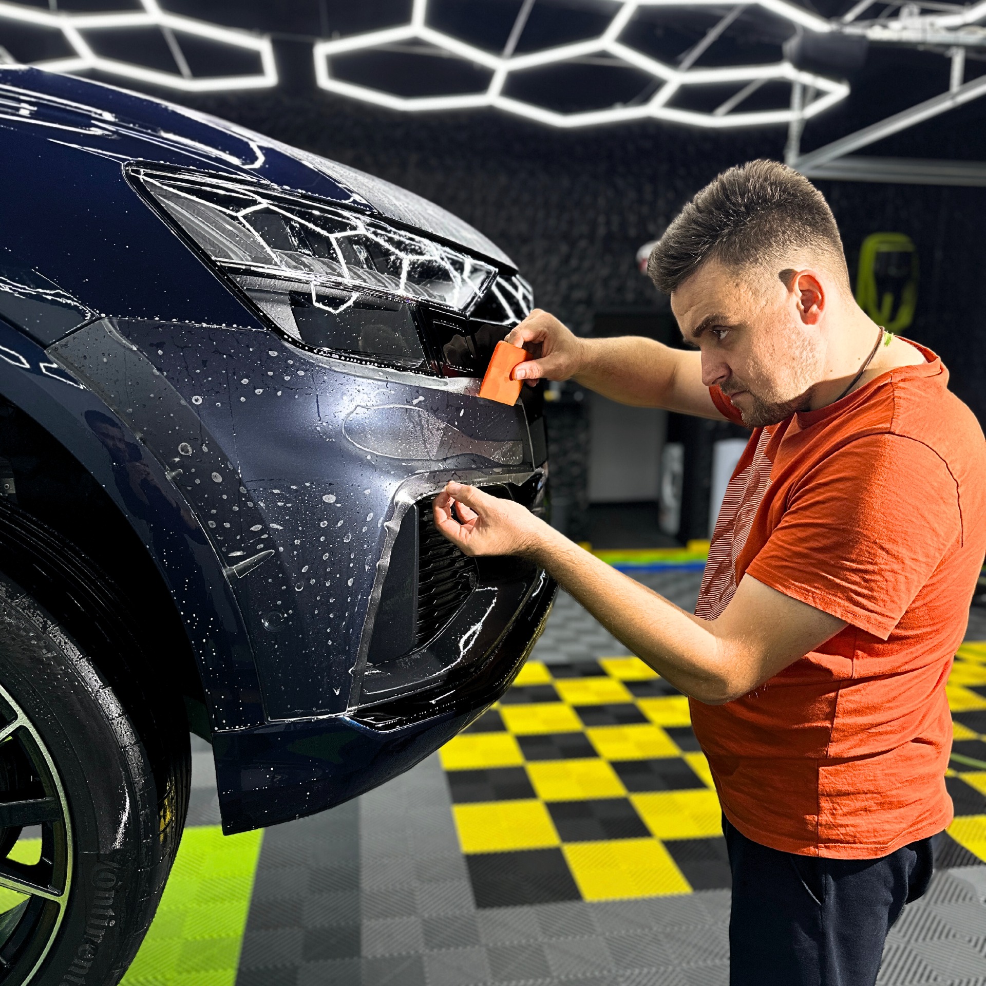 Aplikacja folii ochronnej na przedni zderzak granatowego samochodu w studiu detailingowym, widoczna osoba używająca pomarańczowej rakli do wygładzania folii, w tle podłoga w szachownicę...