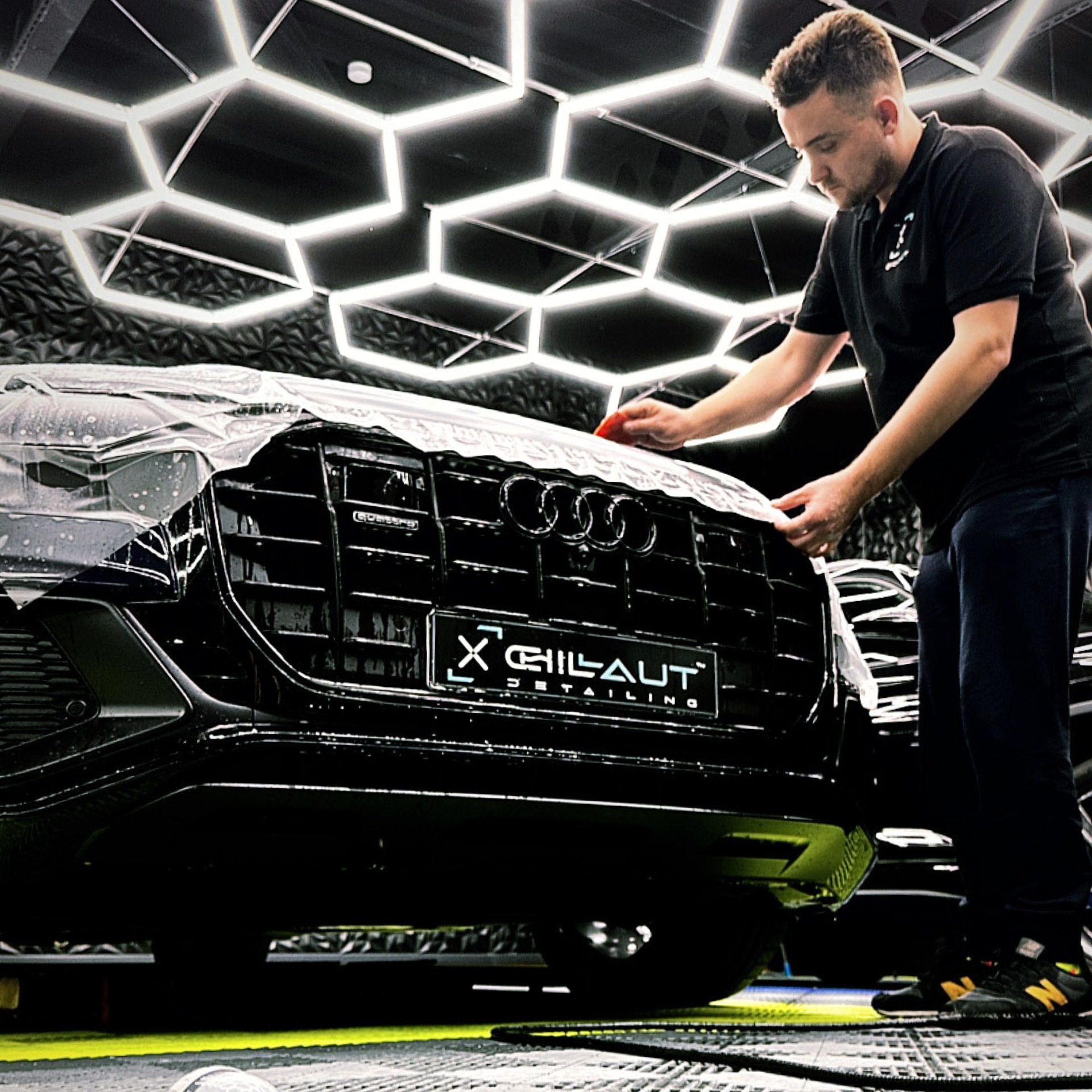Pracownik w czarnym polo nakłada folię ochronną na przedni grill czarnego samochodu Audi w studiu detailingowym z oświetleniem LED w kształcie plastra miodu na suficie.