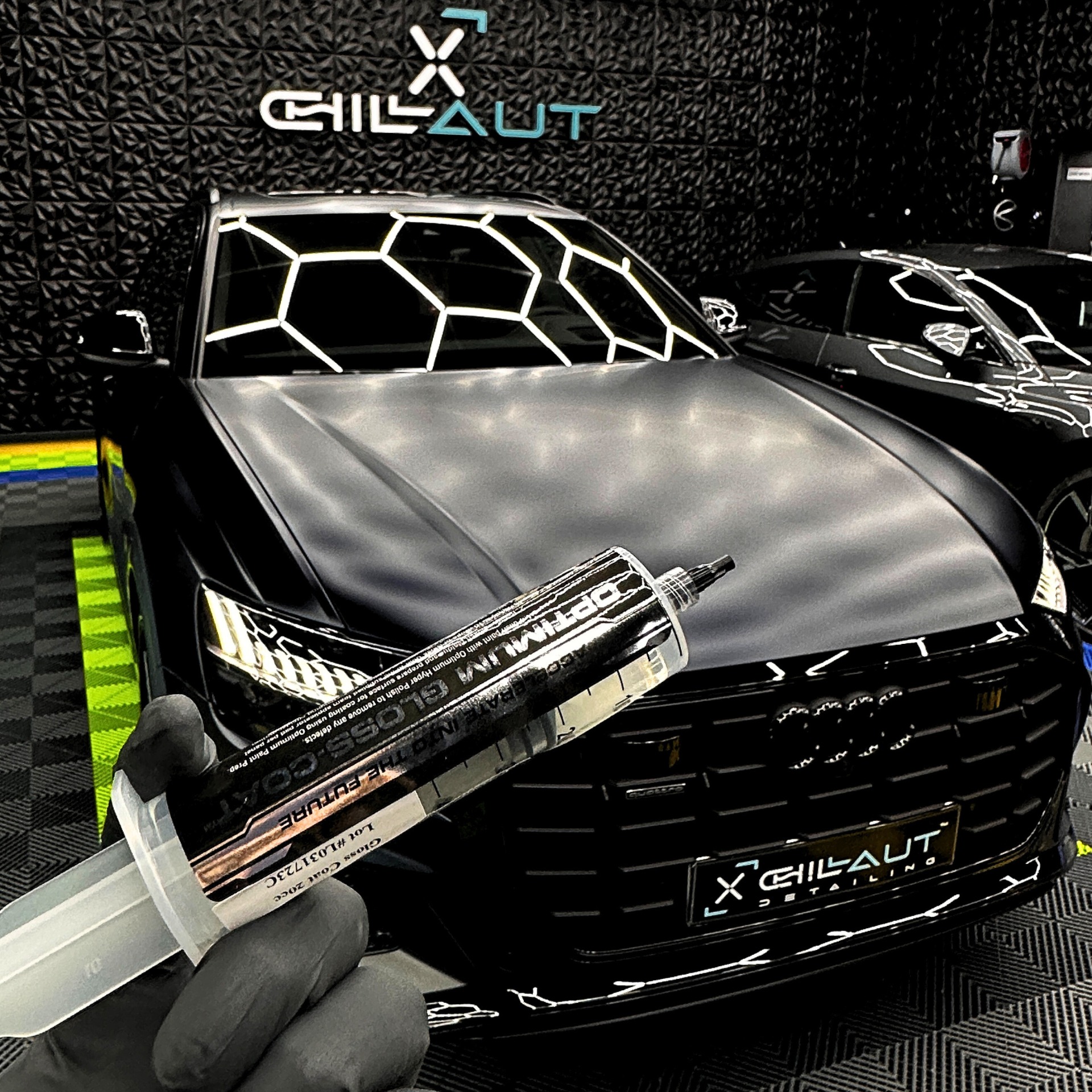 Aplikacja powłoki ochronnej na matowy, czarny samochód Audi, z użyciem preparatu Optimum Gloss-Coat w strzykawce, w tle logo firmy detailingowej na teksturowanej ścianie.