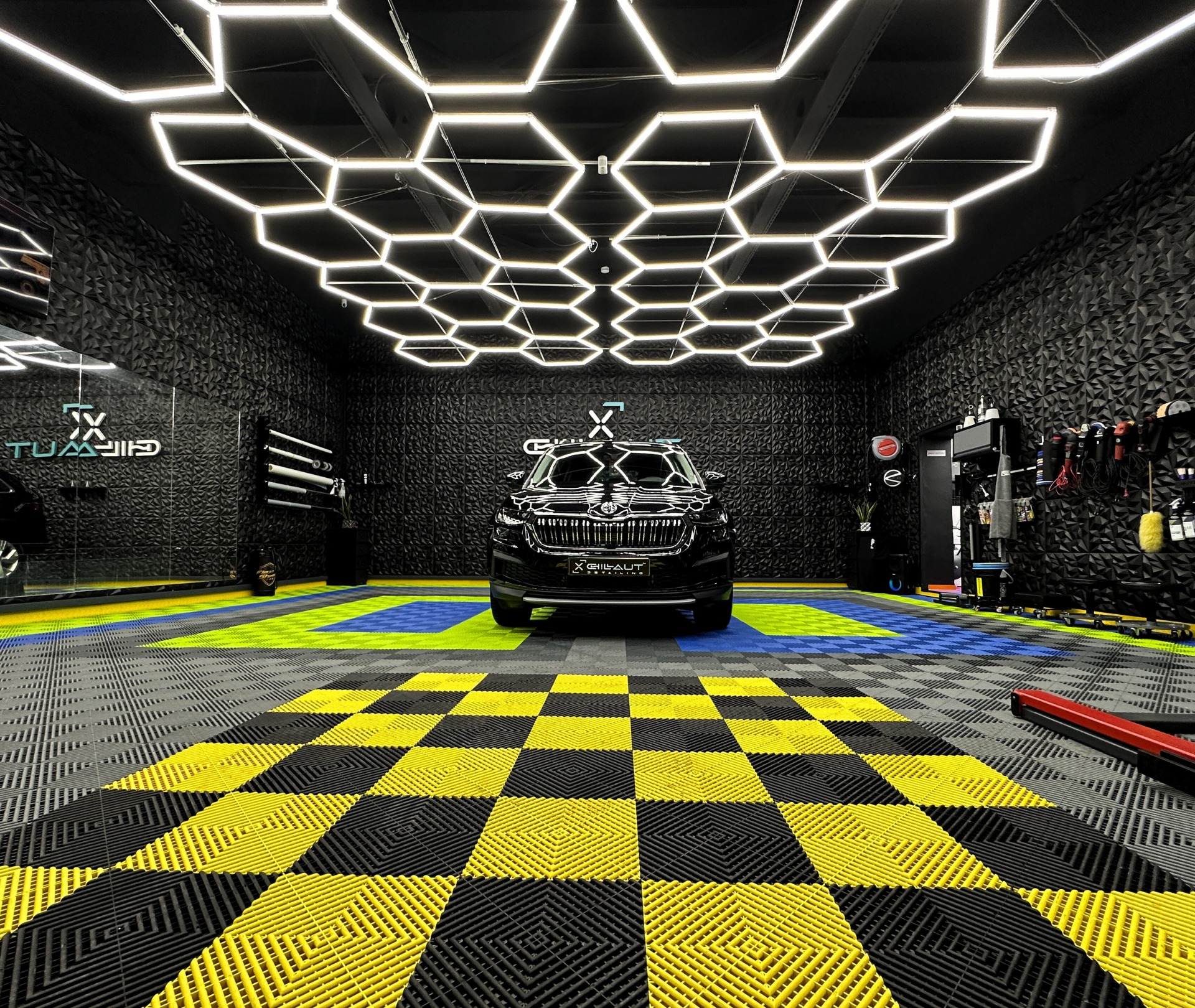 Czarne auto w studiu detailingowym z heksagonalnym oświetleniem sufitowym i podłogą w szachownicę.