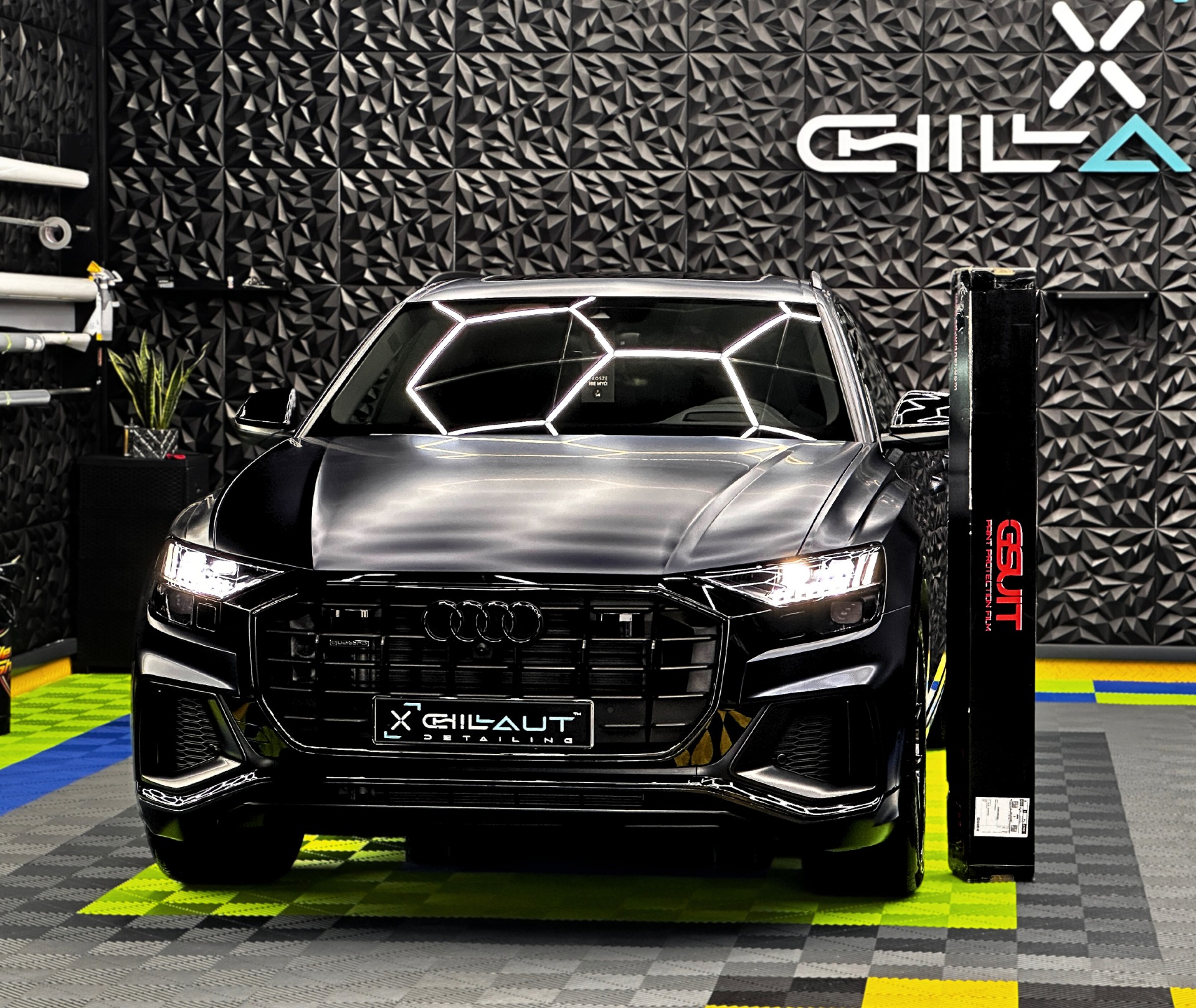 Czarne Audi Q8 po detailingu, z folią ochronną Gsuit, na podłodze w szachownicę, z logo Gilaut Detailing na grillu i dekoracyjną ścianą 3D w tle.