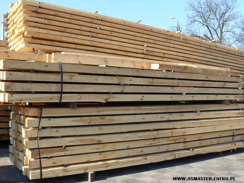 Stosy świeżo przetartego drewna konstrukcyjnego, ułożone na paletach i zabezpieczone czarną taśmą, widoczne na zewnątrz w słoneczny dzień.