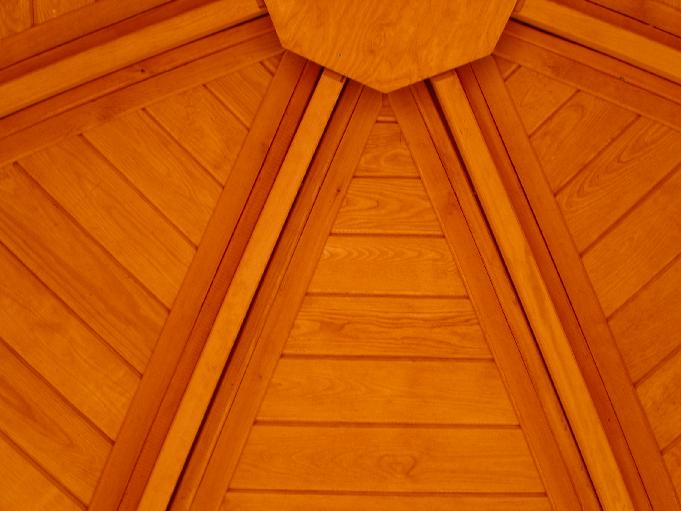 Drewniany sufit altany ogrodowej z widoczną konstrukcją nośną i deskami ułożonymi pod kątem, w ciepłym odcieniu brązu.