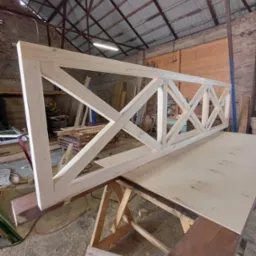 Surowa, drewniana rama z krzyżowymi wzmocnieniami, prawdopodobnie fragment balustrady, ułożona na stole warsztatowym w hali produkcyjnej.