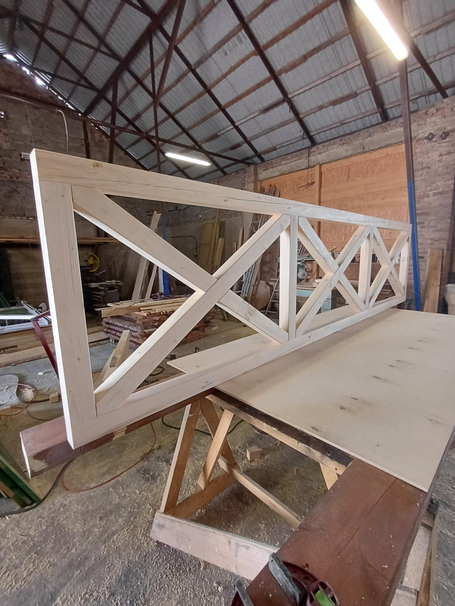 Surowa, drewniana rama z krzyżowymi wzmocnieniami, prawdopodobnie fragment balustrady, ułożona na stole warsztatowym w hali produkcyjnej.