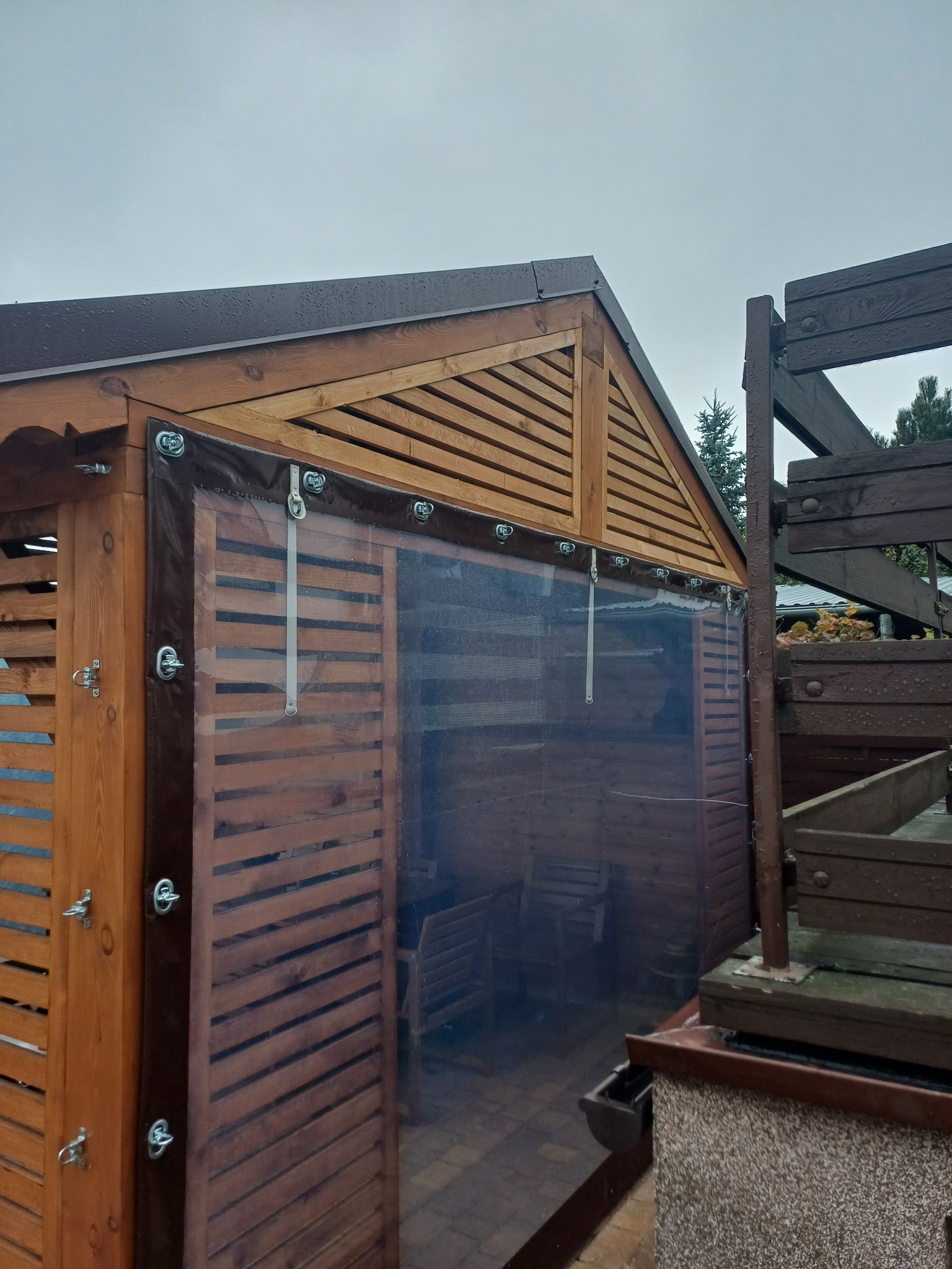 Drewniana konstrukcja z zasłoniętą częścią przezroczystą folią, mocowaną na metalowe zaczepy, widoczne drewniane elementy dekoracyjne i zadaszenie.