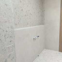 Układanie Płytek Rafał Szczepański - Jasnoszara łazienka z płytkami imitującymi kamień, widoczne przyłącza wodne i odpływ liniowy. Minimalistyczny design, neutralna kolorystyka.