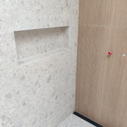 Układanie Płytek Rafał Szczepański - Wykończona łazienka z wnęką w ścianie wyłożonej szarymi płytkami lastryko i ścianą z płytek imitujących drewniane lamele. Widoczny fragment podłogi i czarny odpływ liniowy.